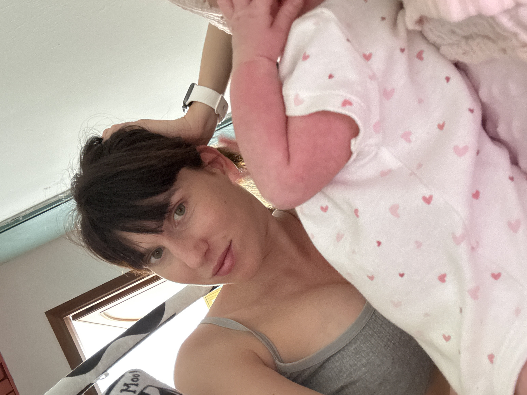 This intimate, upward-angled shot features a mother and her baby indoors in Isola delle Femmine, Italy, during daytime. The woman, with dark hair pulled back and wearing a gray tank top, looks directly at the camera with calm, green eyes and a slight smile. A white smartwatch is visible on her left wrist, and her right arm is raised, perhaps adjusting her hair or supporting the baby.
Partially visible in the upper right is the baby, dressed in white clothing patterned with numerous small pink hearts. The baby's delicate, pink-toned arm and a small portion of its face, obscured by its hand, are visible. Soft pink fabric or a blanket is also present near the baby.
Natural light floods the scene from a window or skylight in the background, hinting at bright daylight. A dark brown window frame is visible, along with a glimpse of a light blue-green element. In the lower left foreground, a black and white striped fabric and a white tag with the word "Moo" printed on it are discernible. The image conveys a tender, casual moment of motherhood.