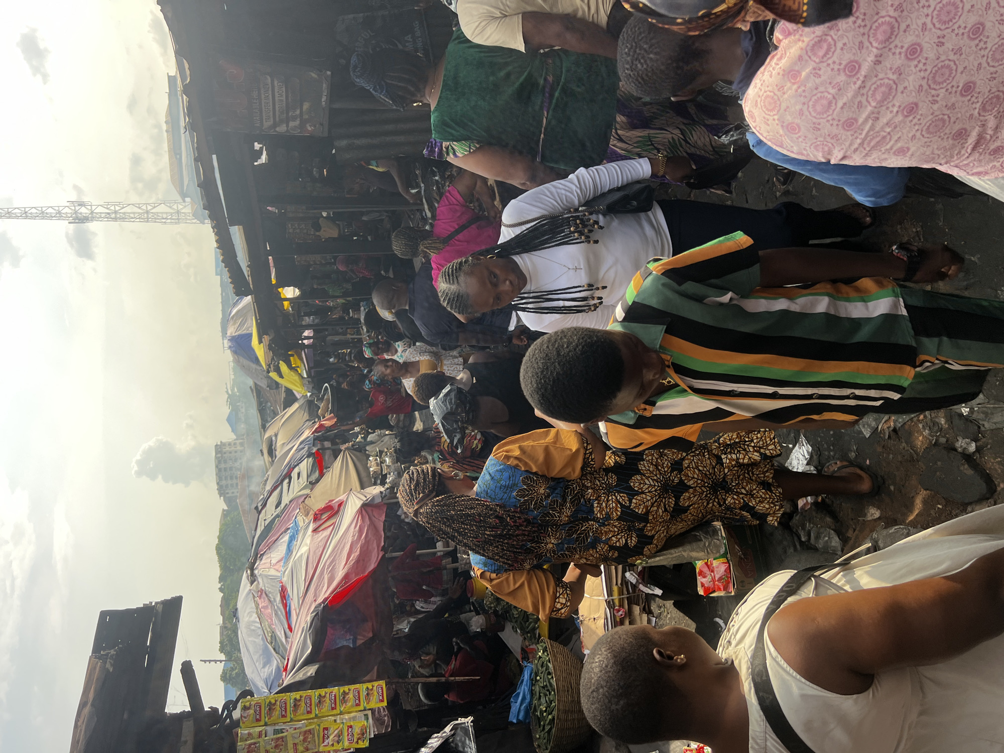 In Onitsha, Nigeria, a bustling market teems with activity under a hazy sky. The scene is packed with people, many of whom are women wearing vibrant clothing, navigating narrow pathways lined with stalls. Some stalls are covered with tarps and umbrellas, displaying goods such as food items in yellow packets and baskets of green produce. In the background, a tall telecommunications tower rises above the makeshift structures, and a plume of smoke ascends into the sky. The ground is muddy and uneven, suggesting an outdoor, unpaved area. The atmosphere appears busy and chaotic, characteristic of a busy marketplace.
