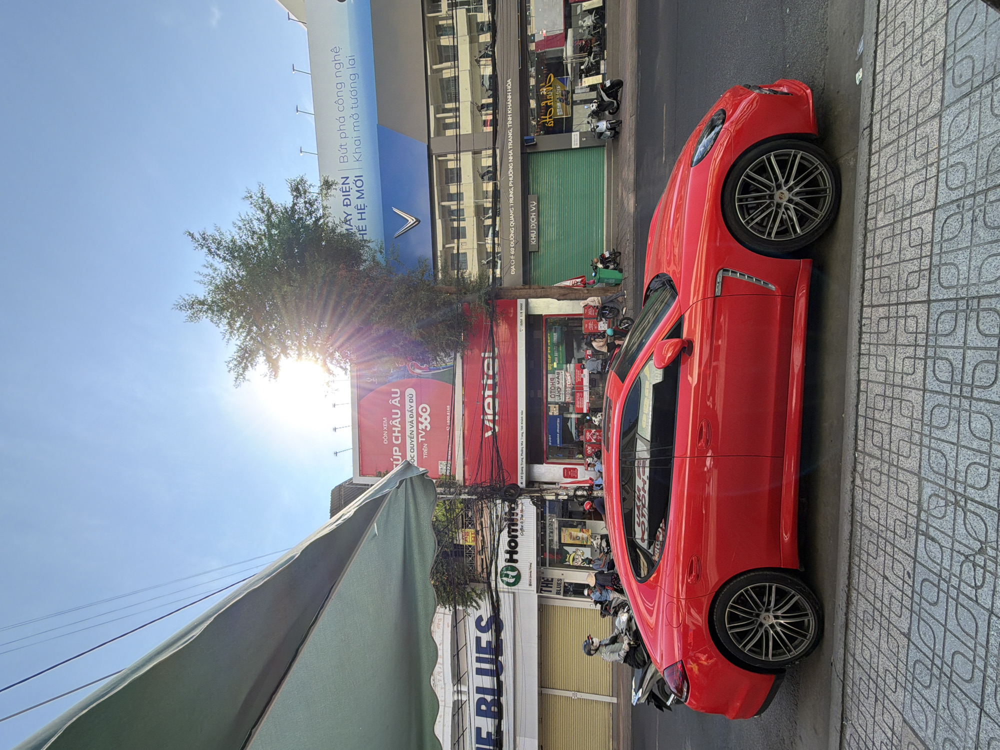 A bright red luxury sedan is parked along a sidewalk in Nha Trang, Vietnam. The car, likely a Porsche Panamera, occupies the foreground and mid-ground. Behind the car, a busy street is visible with various shops and billboards. A green awning extends from a building on the left side of the image. Several people on motorbikes are present on the street, some interacting with shopkeepers. Buildings display advertisements in Vietnamese. A large billboard for VinFast electric vehicles is prominent in the background, along with other commercial signage. The sky is clear and blue, with the sun shining brightly, casting lens flares. The sidewalk is paved with a decorative pattern of grey tiles.