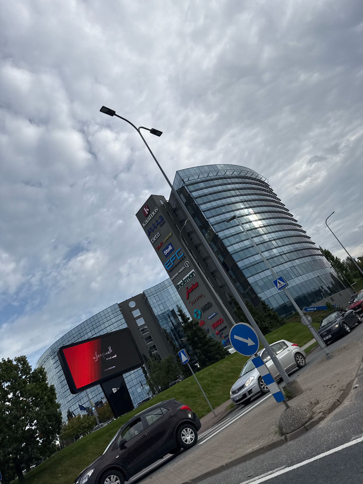 This is a wide-angle, eye-level, slightly tilted photograph of a modern office building complex in Tallinn, Estonia, on a cloudy day. The building features a curved, glass facade that reflects the sky. In the foreground, there is a street with several cars, including a dark-colored hatchback, a silver sedan, and a black convertible. The cars are parked or driving on the road and sidewalk. A tall signpost displays the logos of various companies such as "kaamos," "tieto," "Stell," and "EFC." To the left of the building, a large digital billboard displays the word "Sleepwell" with red and black graphics. The scene also includes streetlights, trees, and a pedestrian crossing sign. The overall impression is a modern urban environment with commercial activity.