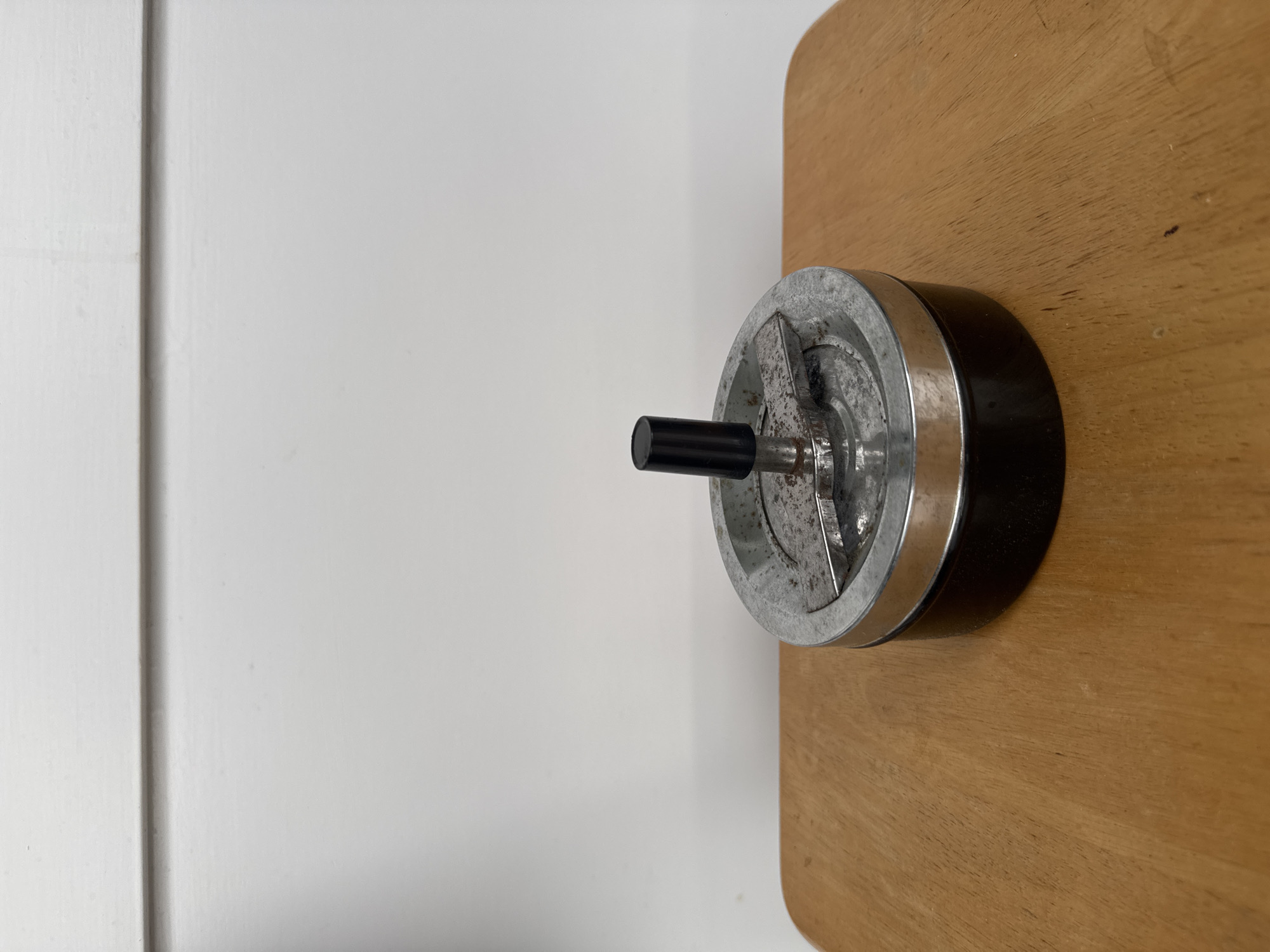 The image shows a close-up, eye-level shot of a vintage ashtray sitting on a wooden surface. The ashtray is round with a silver metal lid and a black base. The lid has a spinning mechanism in the center with a black handle and a metal arm that can be used to extinguish cigarettes. The ashtray appears to be used, with some ash and debris visible on the lid. The wooden surface is light brown and has a visible grain pattern. In the background, there is a plain white wall with a horizontal molding near the top. The lighting is even and diffused, casting no strong shadows. The scene is static and devoid of any human presence or activity. The context is an indoor setting, likely a tabletop or shelf.