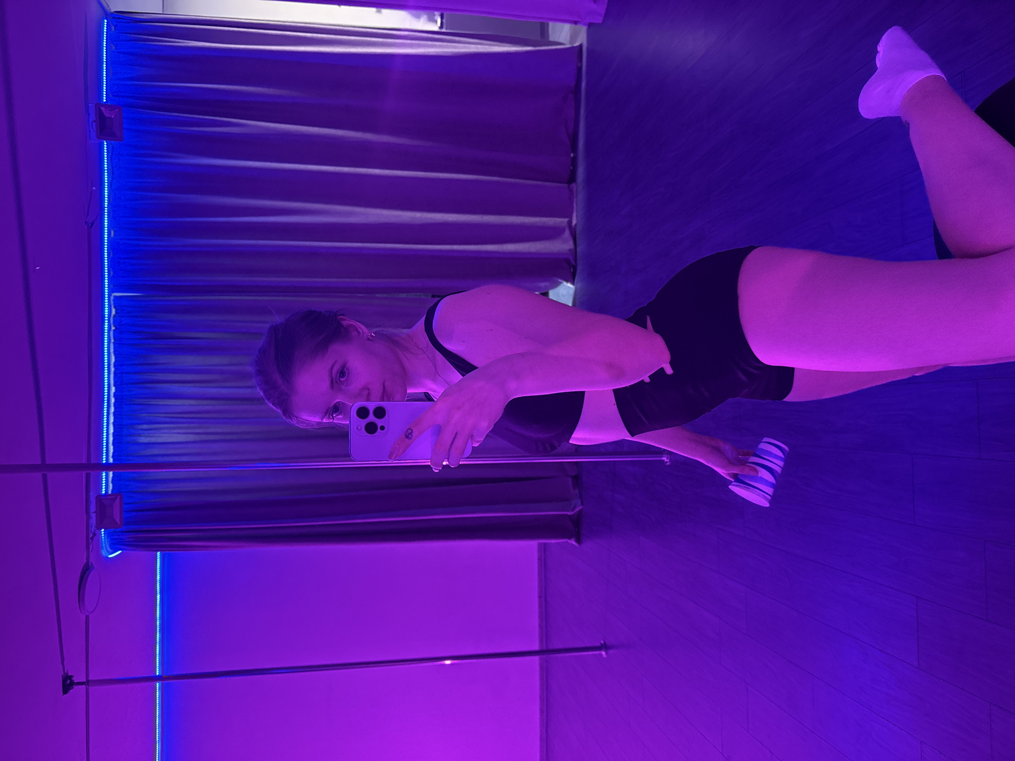 A woman is taking a selfie in a room with purple lighting. She is wearing a black sports bra and black shorts, and white socks on her feet that are visible. She is holding a white phone and a striped cup. There is a dance pole and curtain with string lights behind her. Based on metadata, the image was likely taken in Batumi, Georgia.