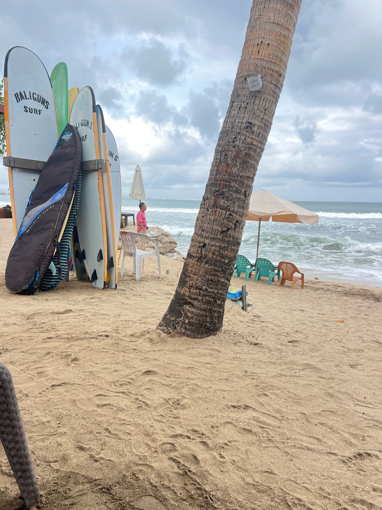 The image shows a beach in Kuta, Indonesia, with a line of surfboards propped up on the sand. The surfboards are of various colors and sizes, with "BALIGUNS SURF" branding visible on some. A black surfboard bag is leaning against one of the boards. In the background, the ocean waves are crashing onto the shore under a cloudy sky. A man in a pink shirt is standing near some sandbags and a white plastic chair. Further down the beach, there are three chairs under a beige umbrella, with the ocean behind them. A large palm tree trunk dominates the foreground on the right side of the image. The sand is a light brown color, with some footprints and debris scattered across it.