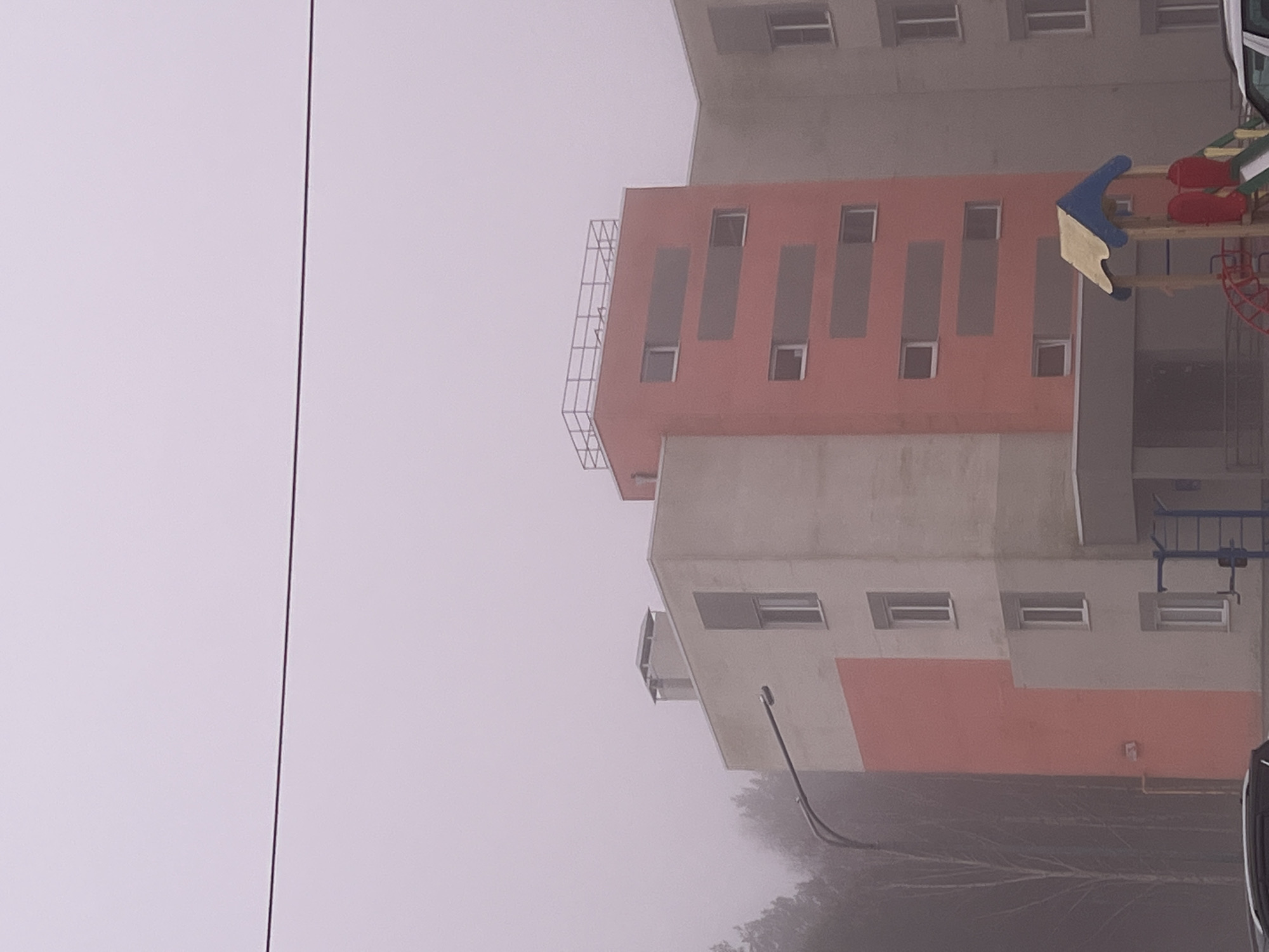 The image shows a multi-story building in Chaikovsky, Russia, viewed from a low angle, possibly from the ground. The building features a combination of concrete and orange-painted sections. There is a playground structure visible on the right side of the building. The scene is obscured by heavy fog or smog, reducing visibility. A power line runs vertically on the left side of the frame. The bottom of the frame shows some trees obscured by the fog. A lamppost is also partially visible.