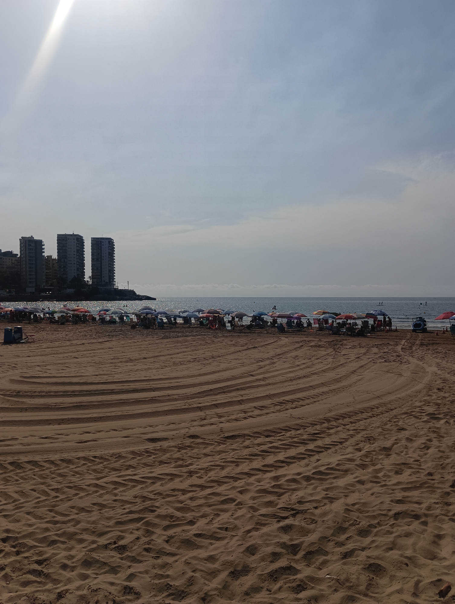 The image depicts a wide sandy beach on a bright, overcast day. In the foreground, the sand shows clear tire tracks, indicating recent grooming or vehicle activity. Further back, a dense row of colorful beach umbrellas and chairs are set up, with scattered beachgoers visible beneath them. Beyond the beach, the calm sea stretches to the horizon, with a few distant figures visible in the water, one appearing to be on a paddleboard. The background features a strip of coastline with several tall, modern residential buildings. The sky is largely overcast with a bright, diffused light source, possibly the sun, creating a glare in the upper left corner. The location is identified as La Playa, Spain.