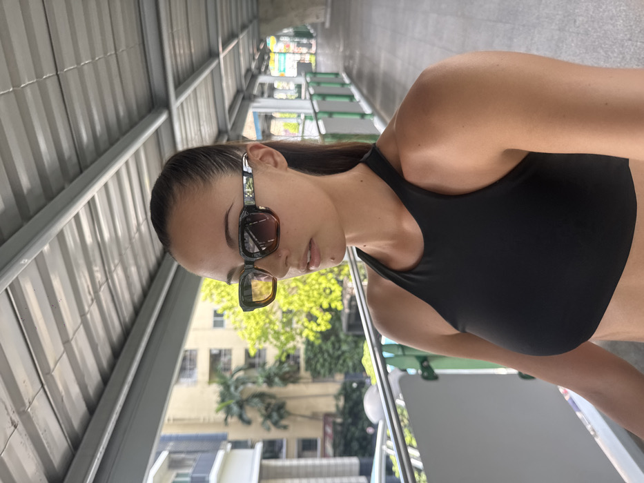 A female individual is depicted standing on an elevated pedestrian walkway, characteristic of the BTS Skytrain system in Bangkok, Thailand. She is positioned slightly off-center to the right, facing towards her left. She wears rectangular sunglasses with a two-tone frame, transitioning from dark to amber, and gradient-tinted lenses. Her dark hair is pulled back, and a small mole is visible below her lower lip. She is dressed in a black, high-neck sports bra. The walkway features a grey, corrugated metal roof, a light-colored paved surface, and a railing with a green horizontal bar on the left side of the frame. In the background, urban multi-story buildings and lush green tropical foliage are visible under bright daylight conditions. The walkway extends into the distance to the right, showing further structural elements of the elevated infrastructure.