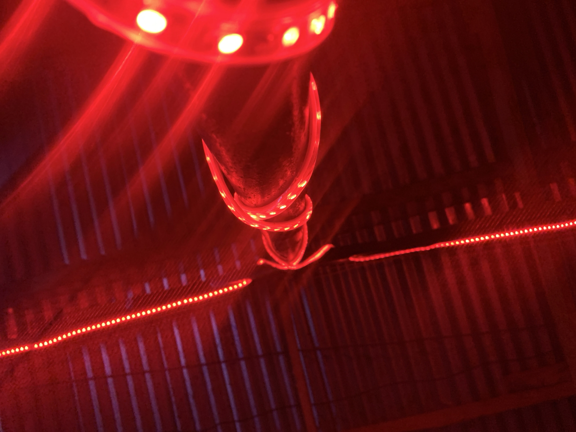 The image depicts an indoor scene, likely a ceiling, illuminated predominantly in red light. The ceiling appears to be constructed of corrugated metal. There are LED strip lights running along the metal, casting a red glow and defining the ceiling's structure. Above, a round light fixture with multiple LED bulbs adds to the overall red illumination. In the center, a spiraled object also lit with LEDs hangs down, creating a focal point. No people are visible, but the scene is bathed in an intense, artificial red light. The location, Kabong, Nigeria, is not evident from the visual elements.
