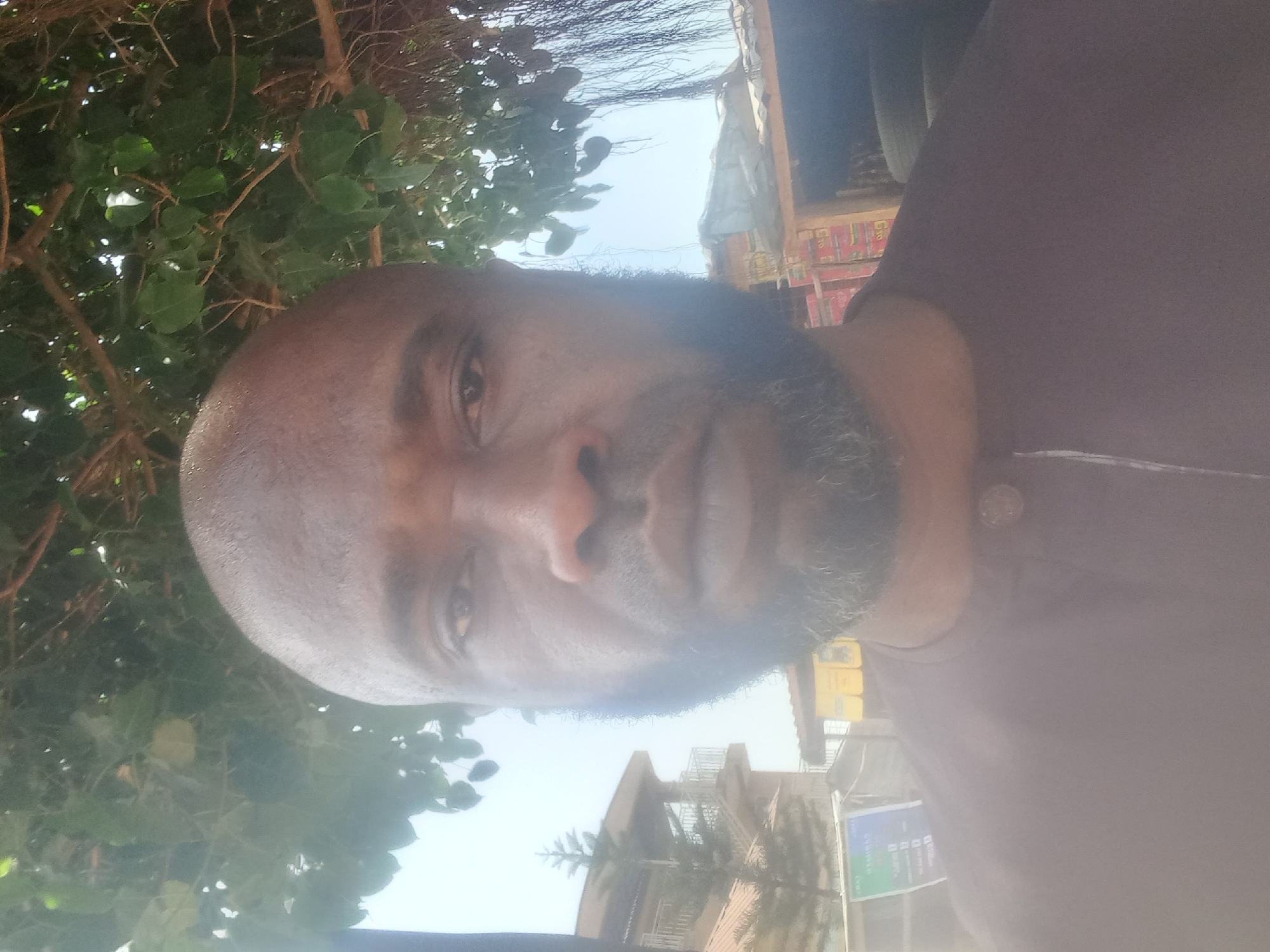 A bald, middle-aged Black man with a graying beard stands near a building with palm trees. He is facing the camera and wearing a dark purple shirt. The background includes buildings, trees, and a partly cloudy sky. The location appears to be in an urban area, possibly Barden Yana, Nigeria, based on the prompt.