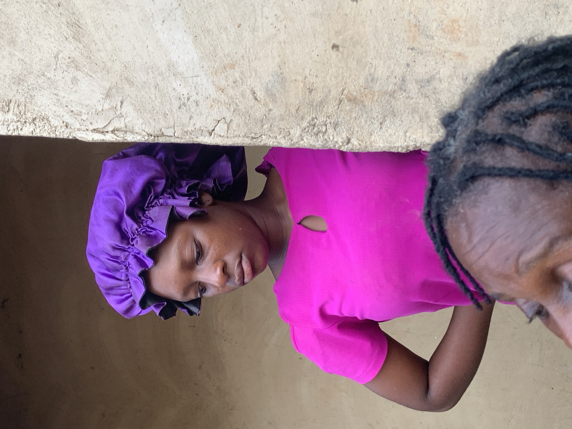The image shows two individuals standing near a building in what is presumed to be Agban, Nigeria. A woman is partially visible on the left side, wearing a purple bonnet and a bright pink shirt with a distinctive keyhole neckline. Her expression is neutral, and she appears to be in her late teens to early twenties. The second individual, on the right side of the frame, is an older adult with tightly braided or loc'd hair. Only a portion of their face is visible. The setting suggests an outdoor environment, likely a residential area, with a concrete or stucco wall running along the top of the frame. The overall ambiance is casual and intimate, as if the photo were taken during a spontaneous interaction.