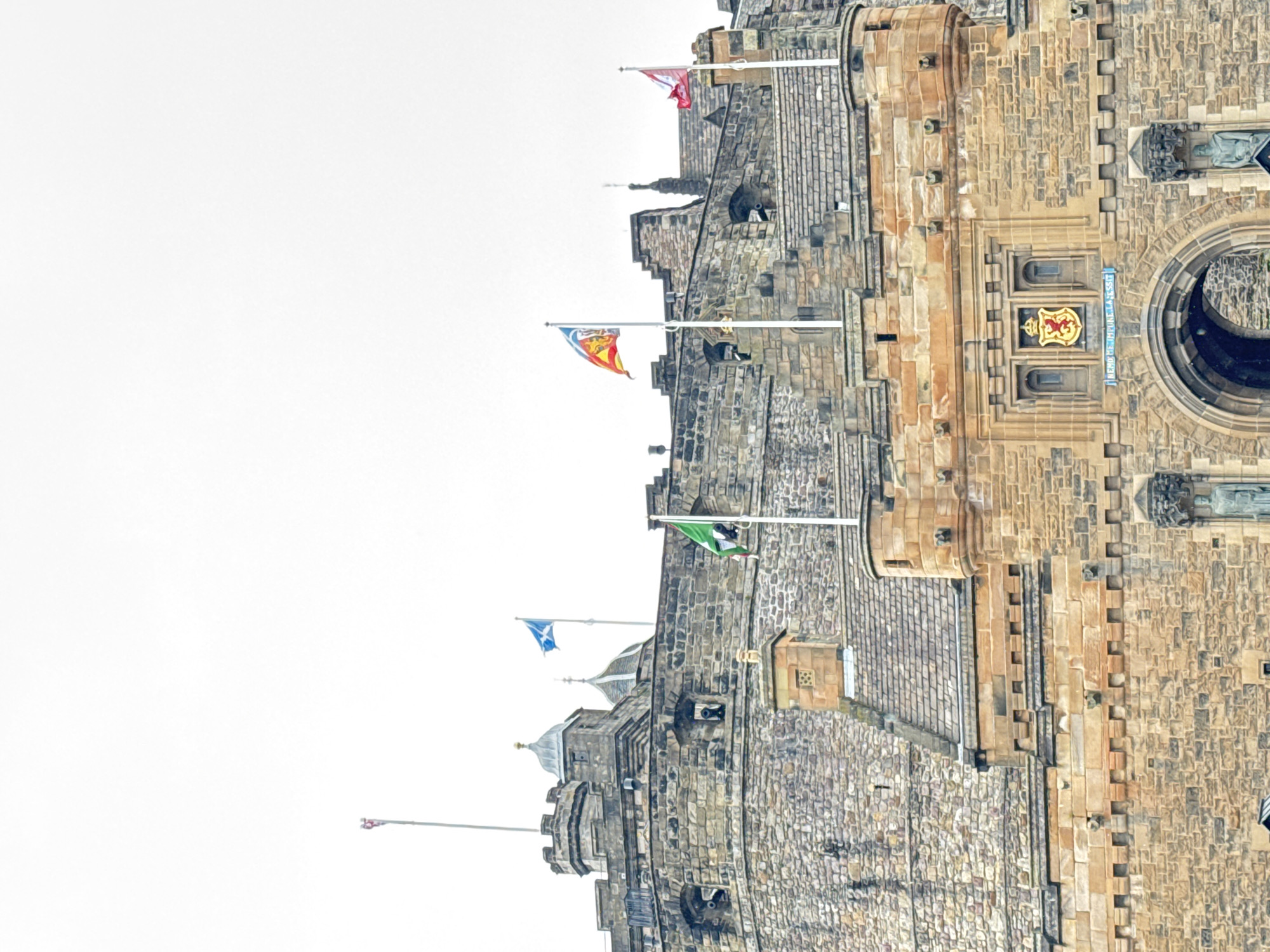 This image shows a low-angle, full-frame view of Edinburgh Castle, located in the city of Edinburgh, United Kingdom. The castle is constructed primarily of stone, with visible turrets, battlements, and architectural details. Several flags are flying from poles on the castle walls. One flag, on the left, is blue with a white diagonal cross, representing the Scottish flag. To its right, a multi-colored flag with red, yellow, and blue elements is visible. Further to the right, a red and white flag is flying. A green flag with a crest is also prominent on the left side of the image. Below the flags and in the central portion of the image, a large stone archway is visible, leading into the castle. Above the archway, a shield with a red lion rampant on a gold background is displayed. A Latin inscription, "NEMO ME IMPUNE LACESSIT," is visible below the shield. The sky is overcast and white, suggesting a cloudy day. The overall scene depicts a historical landmark under a subdued sky.