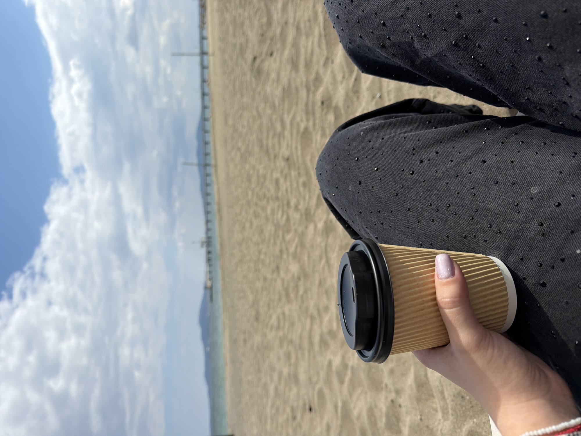 A person holds a disposable coffee cup on a beach near Burgas, Bulgaria. The person is wearing dark pants with small, shiny embellishments. The cup is brown with a black lid. In the background is a sandy beach, the Black Sea, and a distant pier. The sky is partly cloudy.