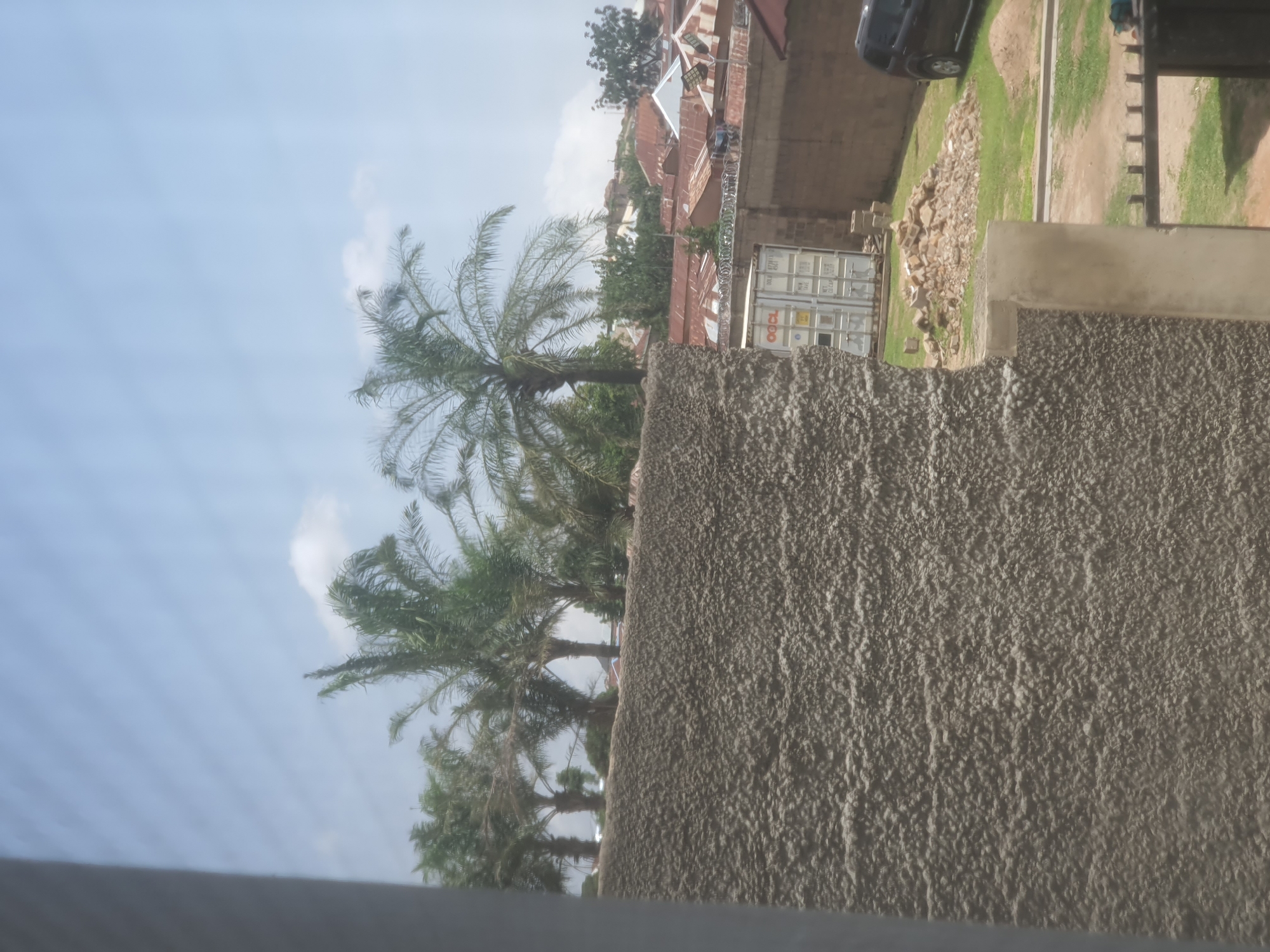 The image depicts an outdoor scene in Jos, Nigeria. The foreground is dominated by a rough, textured wall, likely made of concrete, which takes up the right half of the frame. Beyond this wall, the scene unfolds with several palm trees prominently displayed, their fronds reaching towards a bright, partly cloudy sky. The sky itself suggests daylight, possibly late morning or early afternoon, with a mix of blue sky and scattered white clouds.
In the middle ground, glimpses of buildings with corrugated iron roofs are visible, indicating a residential or commercial area. One notable object is a white shipping container, emblazoned with red text that includes "OCL" and "MAERSK." There are also hints of a vehicle, a dark car, in the upper right portion of the image, parked near a grassy area. The overall setting appears to be a slightly undeveloped or transitional urban environment, with a mix of vegetation, structures, and what looks like a rocky, unpaved area.
The image is oriented vertically, suggesting it was taken with the camera held in portrait mode. There are no discernible people actively engaged in an activity. The weather appears to be pleasant, with the bright sky suggesting sunshine, though the presence of clouds indicates it might not be intensely clear. The combination of palm trees, the style of the buildings, and the overall atmosphere are consistent with the location context of Jos, Nigeria. The notable text visible is on the shipping container: "OCL" and "MAERSK."