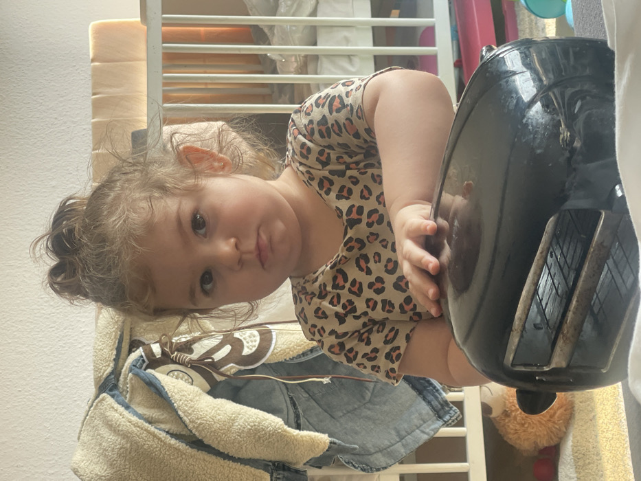 The image captures an indoor scene in Senden, Germany, featuring a toddler-aged child positioned in front of a black toaster oven. The child has light brown, curly hair pulled into a top knot, wears a short-sleeved, leopard-print shirt, and has a small gold earring in the visible right ear. Their expression is a slight pout as they look directly at the camera. The child's right hand rests on the reflective, black top surface of the toaster oven, which displays interior wire racks through its front opening. In the background to the left, a beige-lined denim jacket hangs, partially obscuring a stuffed animal and red toys on a striped rug. To the right, a white wooden crib with light-colored padding is visible. The foreground indicates a soft, light-colored surface, possibly a blanket or rug.