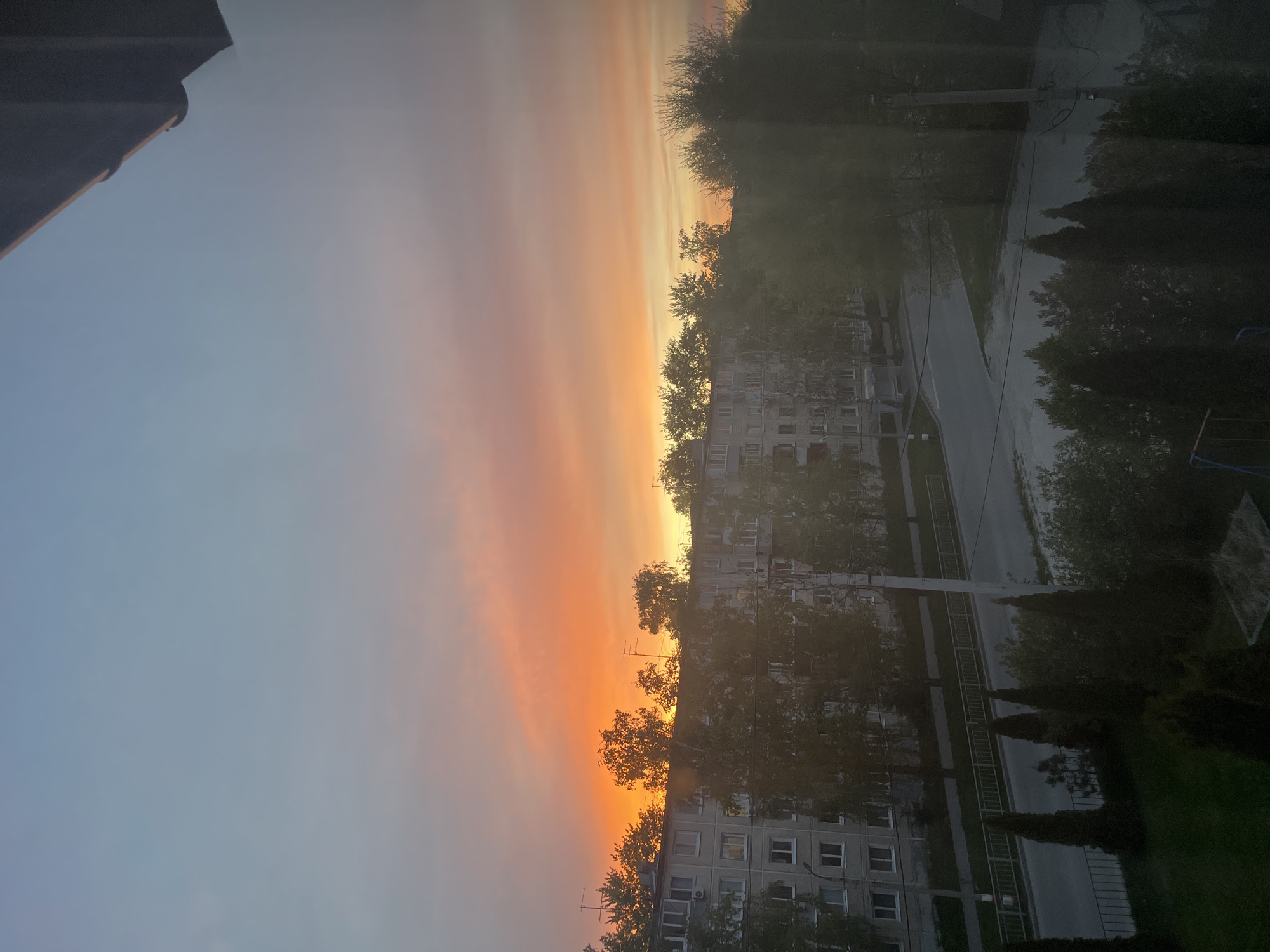 The image shows a cityscape view at sunset. In the foreground are apartment buildings, likely Soviet-era, and some trees. The sky is orange and yellow near the horizon, fading to a pale blue higher up. A wet street is visible to the right of the buildings. The image was likely taken from a high vantage point, possibly another apartment building. Given the architecture, street layout, and presence of Soviet-era apartment buildings, it is likely this photo was taken in an urban residential area of Ulyanovsk, Russia.