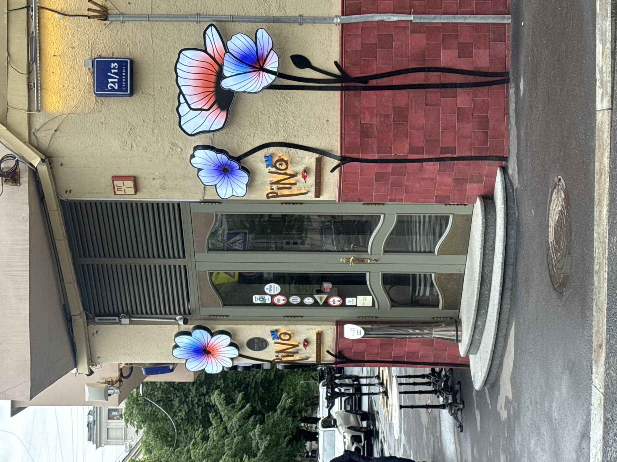 This is an exterior shot of the restaurant "Pino," located in Moscow, Russia. The restaurant's facade features a tan-colored wall with floral decorations, a sign that reads "Pino," and a dark brown door. The address plaque, "21/13," is visible near the top left. Red tiles cover the upper right portion of the wall. A section of gray sidewalk and street is visible on the right. The scene is captured during daylight hours, with outdoor seating visible at the bottom of the image.