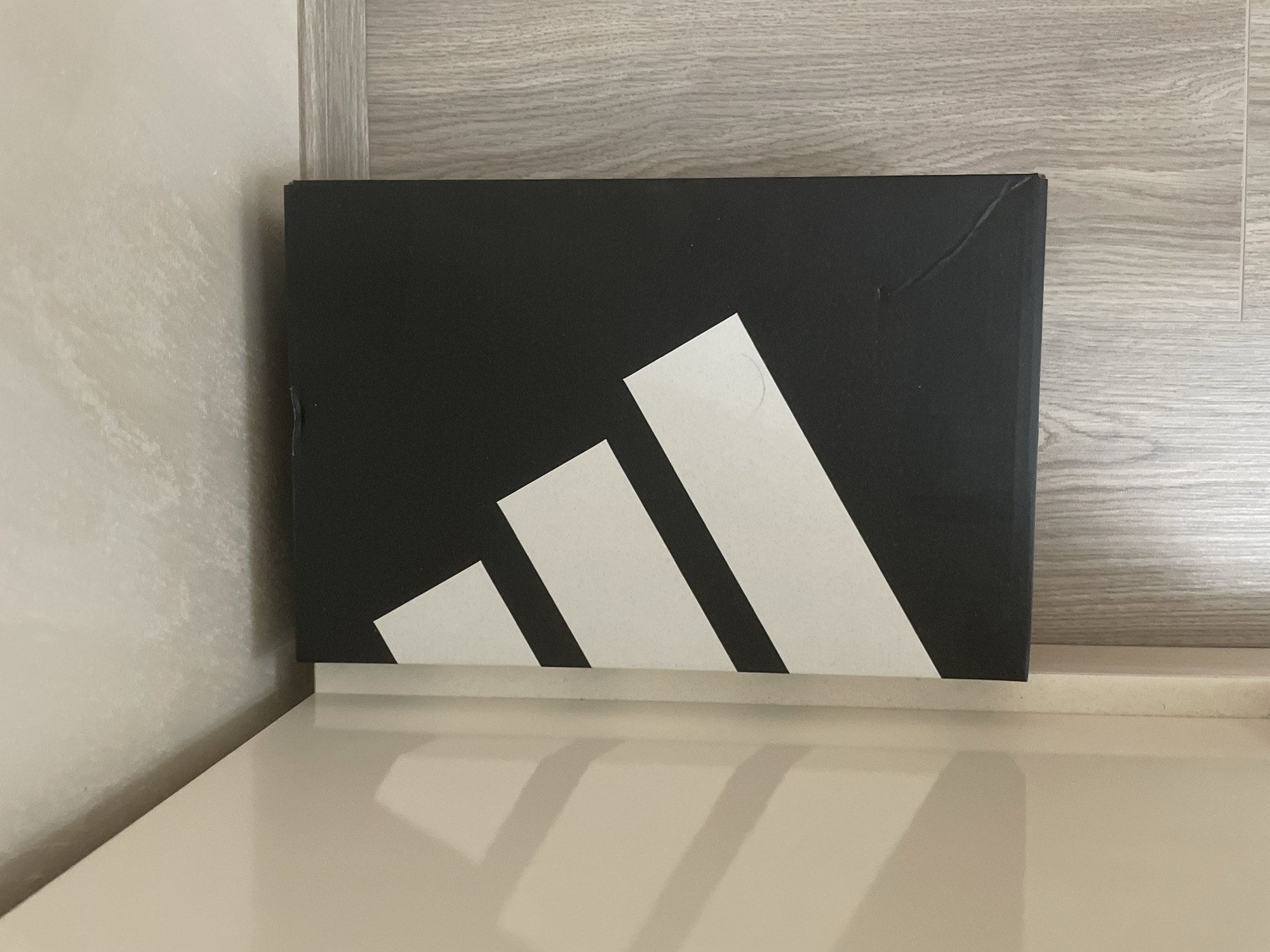 The image captures a black Adidas shoebox, prominently featuring the iconic three white stripes logo. The stripes are angled upwards from left to right across the front of the box. The shoebox rests on a light-colored, reflective surface, possibly a shelf or table, which shows a faint reflection of the white stripes. To the left of the box, a textured, light beige or cream-colored wall is visible, while a light-toned wooden panel with a horizontal grain forms the background directly behind the box.
Several minor imperfections are visible on the shoebox, including a notable crease in the top right corner that extends onto the side. A small scuff mark is present on the left side edge of the box, and a circular impression or mark can be seen on the top rightmost white stripe. The overall lighting is consistent with an indoor setting. No people or activities are depicted in the static shot. The scene is composed of neutral colors, highlighting the black and white design of the Adidas box.