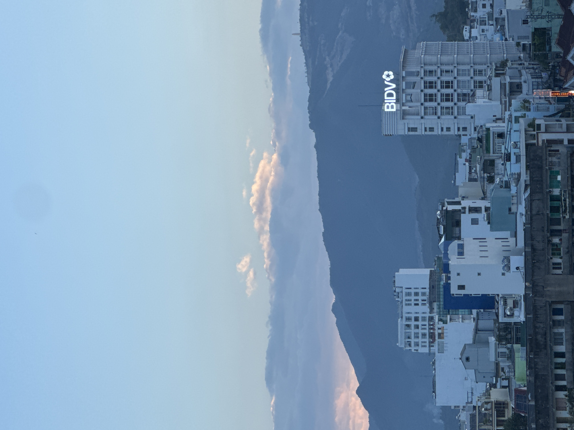 The image is a vertical, eye-level, wide shot of a city skyline in Nha Trang, Vietnam, taken at dusk. The sky is a pale blue with scattered clouds, some tinged pink by the setting sun. In the background, a range of dark blue mountains forms the horizon. The middle ground is filled with a dense collection of urban buildings of varying heights and architectural styles. A prominent white building with the illuminated logo "BIDV" on its rooftop stands out on the right. Other buildings include a tall white structure with many windows on the left, and various smaller residential and commercial buildings in shades of white, blue, and green. Some buildings have visible balconies and window frames. The foreground shows the tops of older, darker buildings. The overall impression is a bustling city nestled against a natural mountain backdrop at the end of the day.