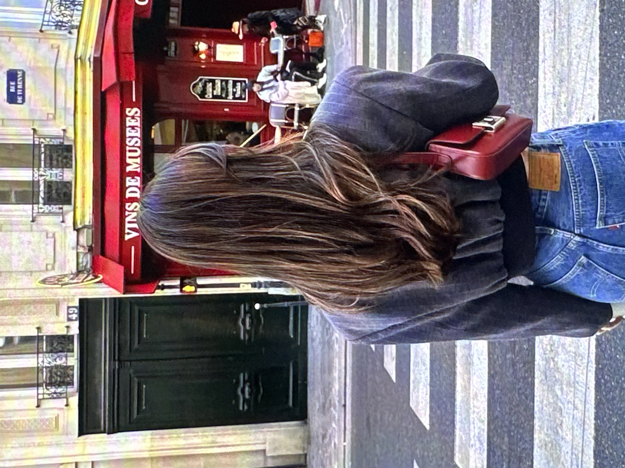 The image shows a person with long brown hair standing on a crosswalk in front of a building. The person is wearing a grey jacket, blue jeans, and a red purse. The building has a red awning with the words "VINS DE MUSEES" written on it. A blue street sign is partially visible, reading "RUE DE TURENNE". The environment appears to be urban, possibly Paris, France, and not Yerevan, Armenia, based on the street sign and building style.