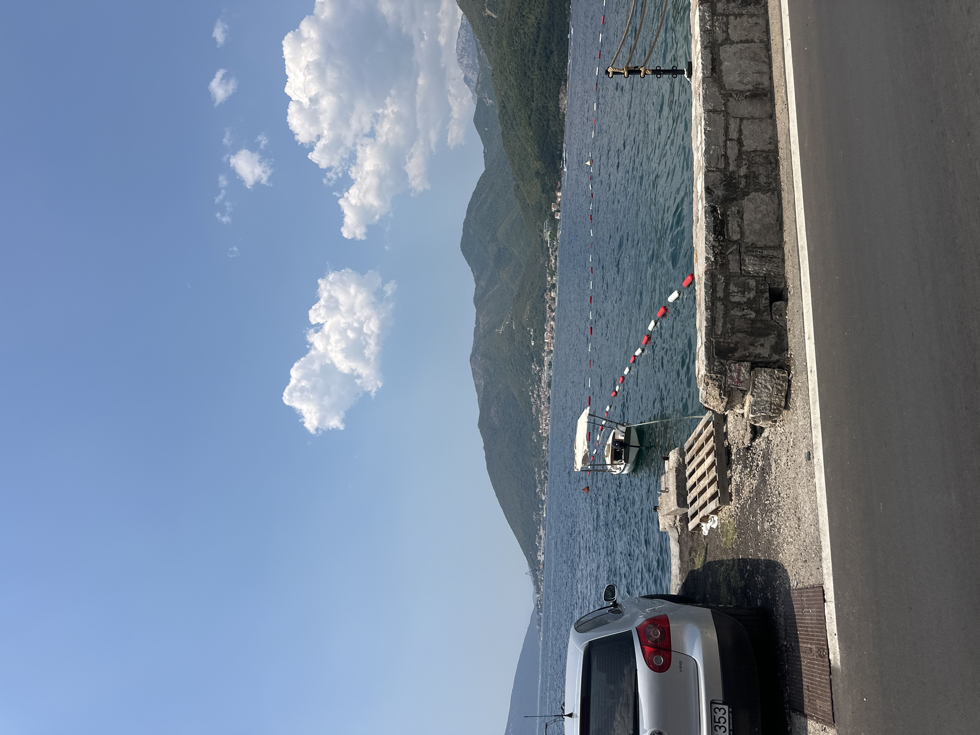 The image shows a view of the Bay of Kotor from the shore in Donja Lastva, Montenegro. In the foreground, a silver Volkswagen Golf car is parked on a paved area next to a stone wall. A wooden pallet lies on the ground near the car. The bay is calm and blue, with a small white boat with a canopy anchored offshore. A line of red and white buoys marks a boundary in the water. In the background, lush green mountains rise from the opposite shore, dotted with buildings and vegetation. The sky is clear blue with a few fluffy white clouds. The overall scene depicts a peaceful coastal setting on a sunny day.