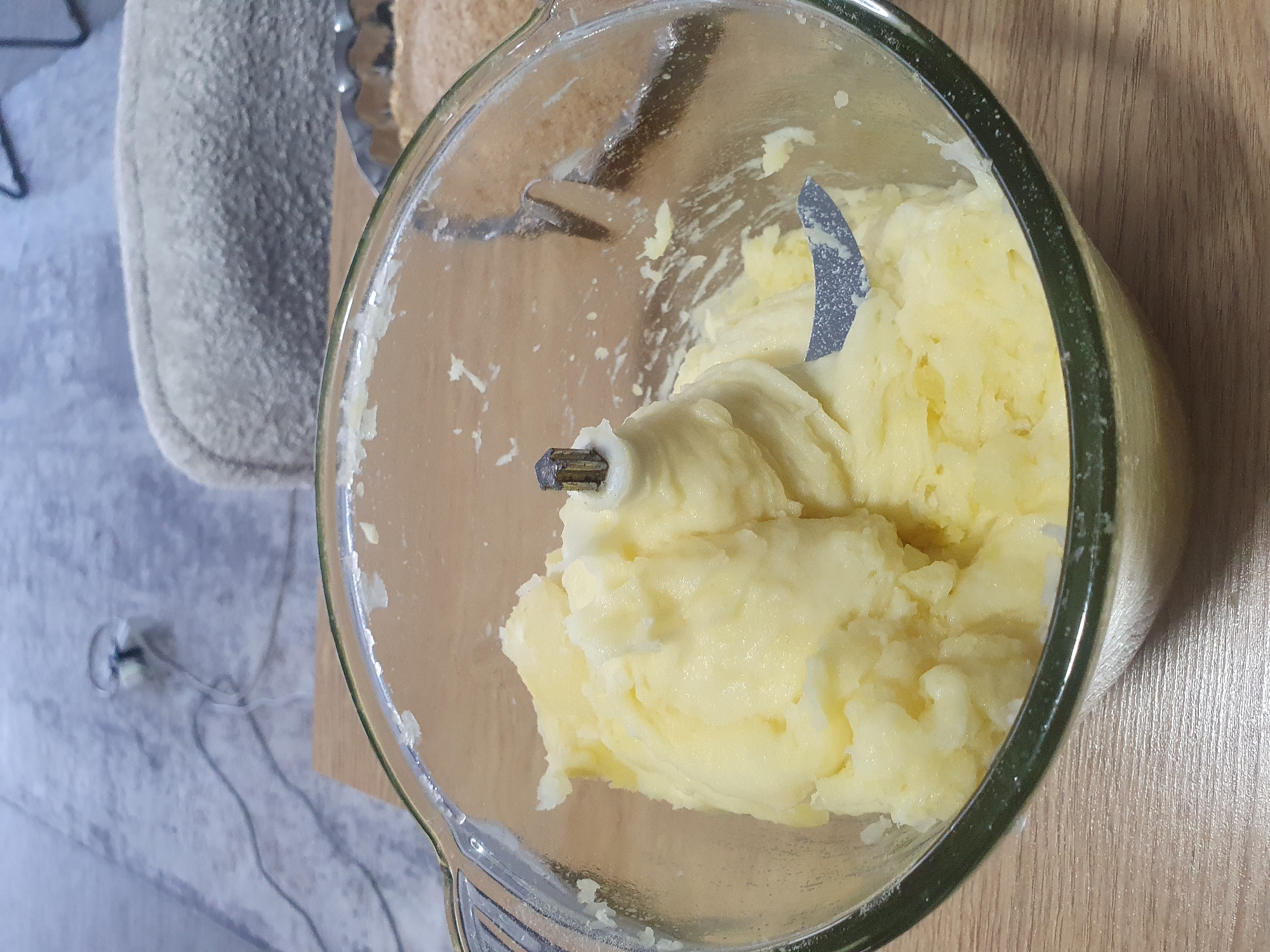 A close-up shot shows mashed potatoes inside a glass food processor bowl. The potatoes are a pale yellow and appear smooth, with some slightly lumpy textures. A metal blade is visible within the bowl, embedded in the mashed potatoes. The bowl rests on a light brown wooden surface. In the background, a blurred gray surface with black cords is visible, possibly a floor with electrical outlets or cables. A lightly textured gray object, possibly a piece of furniture or fabric, is also present. The lighting is warm and somewhat diffused. The scene is indoors. This image is not related to Kheyrabad, Iran.