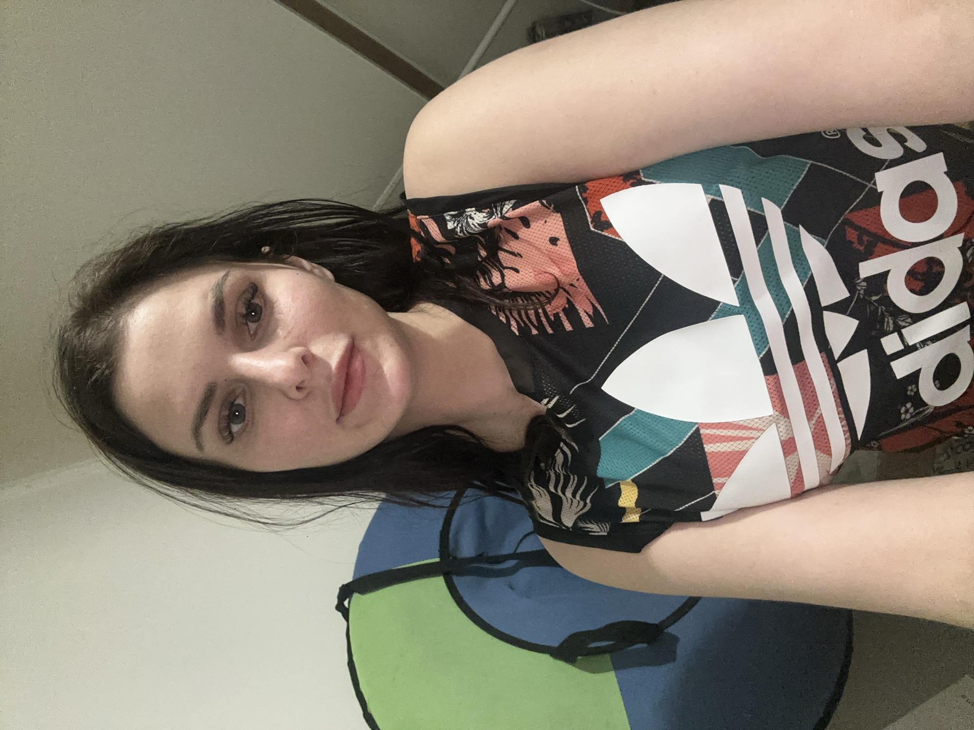 A dark-haired woman is pictured from the chest up, facing the camera. She is wearing a sleeveless black Adidas shirt with a colorful, geometric pattern and a large white Adidas logo. Behind her, to her left, is a large, circular object that is half green and half blue. The background appears to be an indoor setting with a plain wall. The lighting suggests an indoor environment.