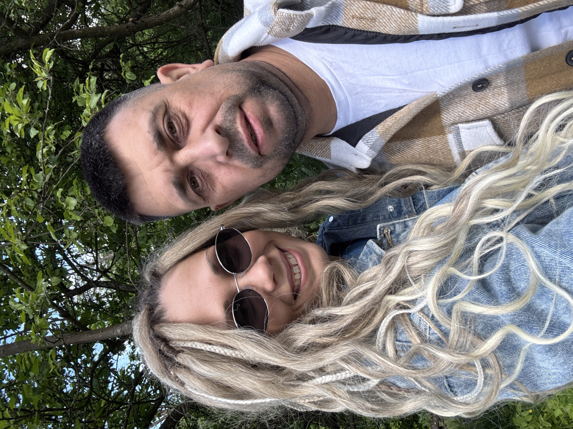 A selfie shows a man and a woman in an outdoor setting, likely in Заволжье, Russia, as indicated in the instructions. The man is at the top of the frame. He has short, dark hair with some graying and stubble. He wears a white t-shirt covered by a brown and white plaid jacket. The woman is below the man. She has long, blonde braided hair and wears round sunglasses and a blue denim jacket. Both individuals are smiling and looking at the camera. Behind them, a background of green trees is visible, suggesting a park or natural setting. The photograph appears to be taken with a phone or similar device.