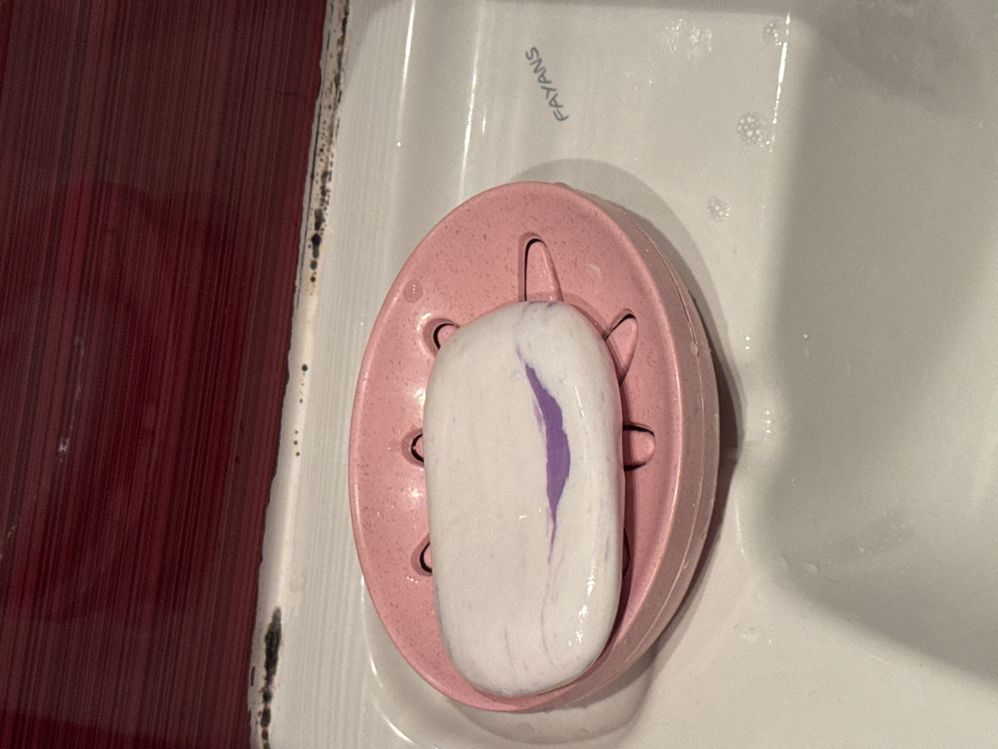 The image depicts a bar of white soap with a streak of purple running through it, sitting on a pink plastic soap dish. The soap dish is located on a white porcelain sink, with water droplets visible on the sink's surface. The sink bears the inscription "FAYANS." To the left of the sink is a red-brown vertical surface with a textured appearance, possibly a wall or cabinet. There is evidence of mold or mildew along the junction between the sink and the red-brown surface.
The image does not provide information to ascertain the location of this scene in Burgas, Bulgaria or any other city.