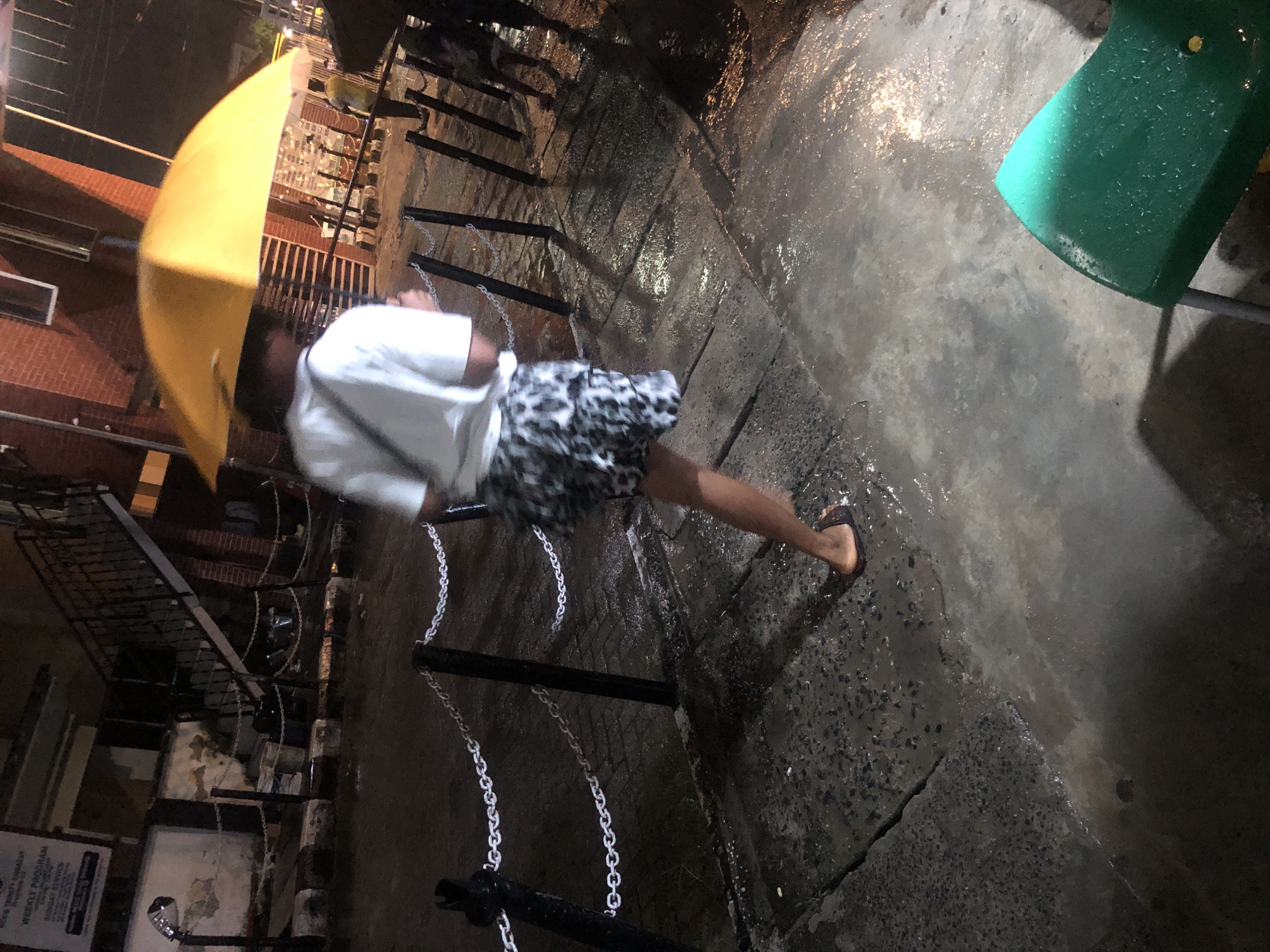 The image depicts a rainy night scene in Ado, Nigeria. A person wearing a white t-shirt and patterned shorts is walking away from the camera, holding a bright yellow umbrella. The ground is wet and reflects the ambient lighting. A chain-link fence with black posts runs horizontally across the mid-ground, separating the pedestrian walkway from a street or building area. In the background, a building with brick details and a staircase is visible. Another person is partially visible in the background, also holding an umbrella. A bright green chair is in the foreground, wet with rain.