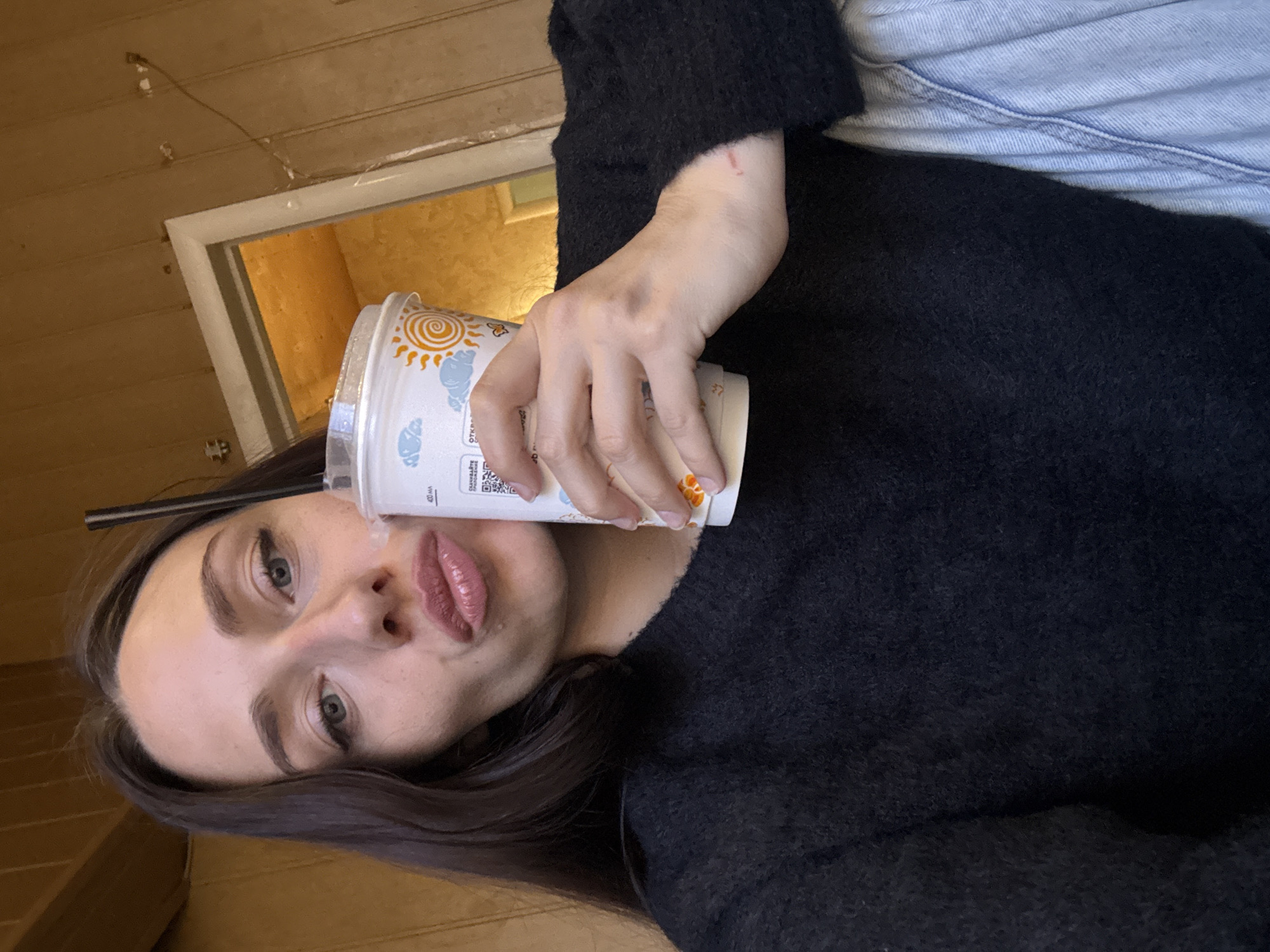 A young woman is holding a disposable cup with an image of a sun and clouds. She is wearing a black sweater and appears to be indoors, possibly in a wooden-paneled room or building. Based on the text visible on the cup, it is likely from the "Teremok" fast-food chain, which has locations in Saint Petersburg, Russia. The woman is holding the cup up towards the camera with her right hand and making a pouting expression. A straw is sticking out of the cup. Light blue denim jeans are visible in the top right corner of the frame.