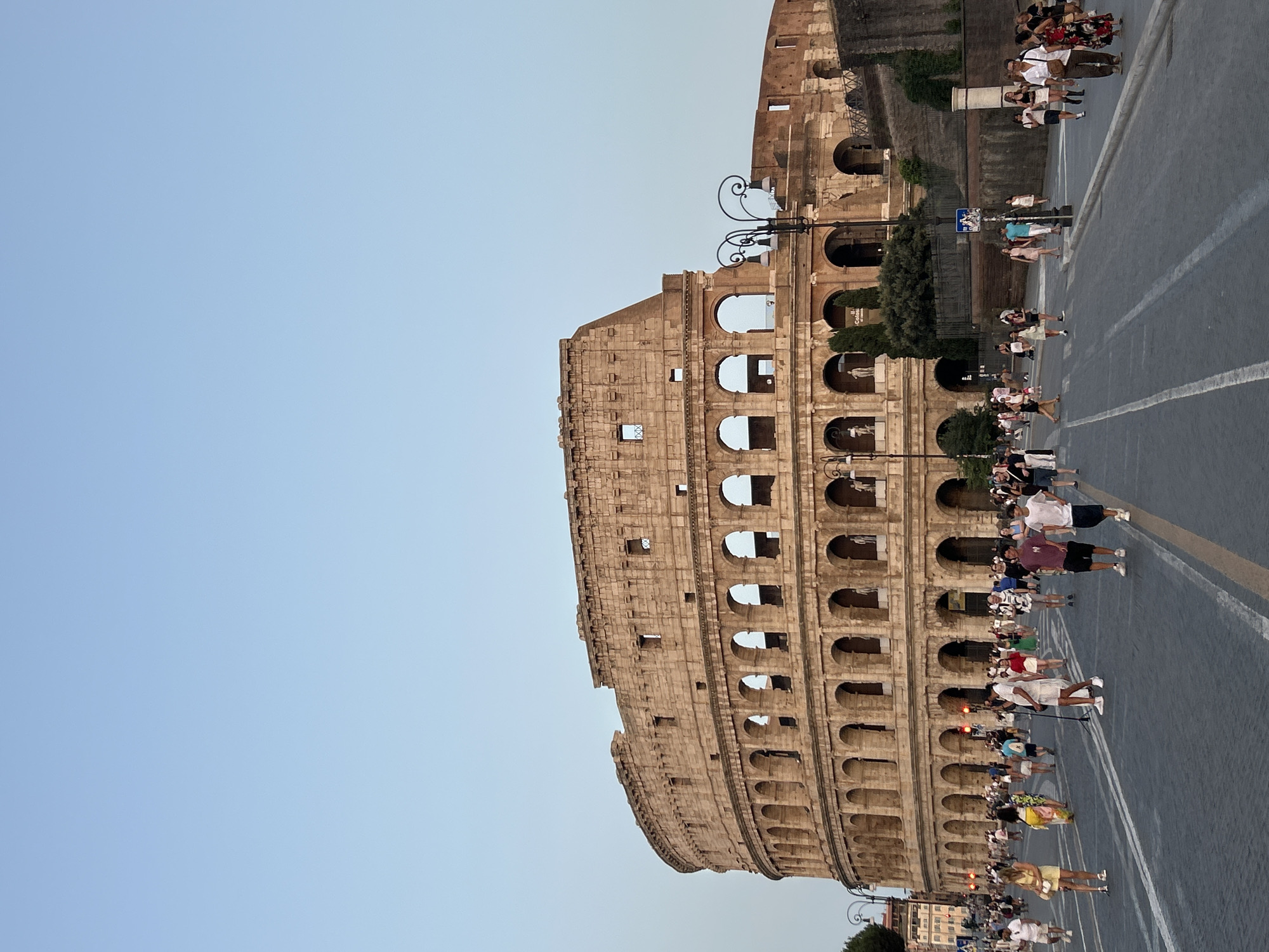 The image shows the Colosseum in Rome, Italy, a large oval amphitheater made of stone. The structure is ancient and shows signs of wear and tear. The sky above is clear and blue, indicating it is daytime. A street runs in front of the Colosseum, with several lanes marked by white lines. Many people are walking on the street and sidewalk, appearing to be tourists. Some are dressed casually in shorts and t-shirts, while others are wearing dresses and more formal attire. There are streetlights and some greenery visible. The overall scene depicts a popular tourist attraction on a clear day, with many visitors taking in the historical monument.