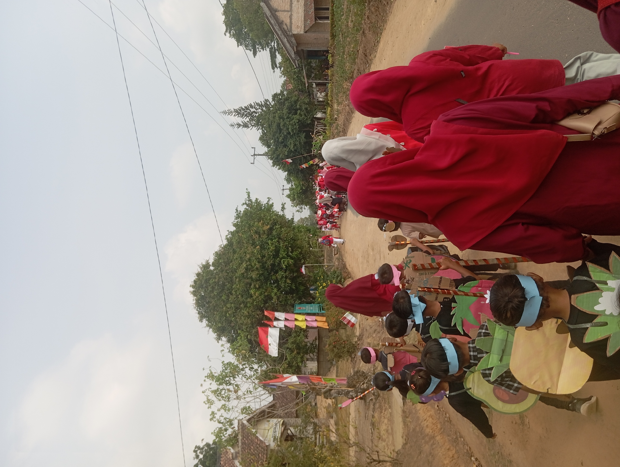 In Dente Teladas, Indonesia, a group of children in a parade march along a dirt road, their backs turned to the camera. They are dressed in various costumes, some with large, fruit-shaped cardboard cutouts attached to their backs, resembling avocados and strawberries. Many of the children are wearing blue headbands. Several adults, mostly women wearing red hijabs, are walking alongside and behind the children. The parade is passing by a rural village with houses and lush green trees. Indonesian flags are visible, suggesting a national celebration or event. The sky is overcast, and the lighting suggests it is daytime.