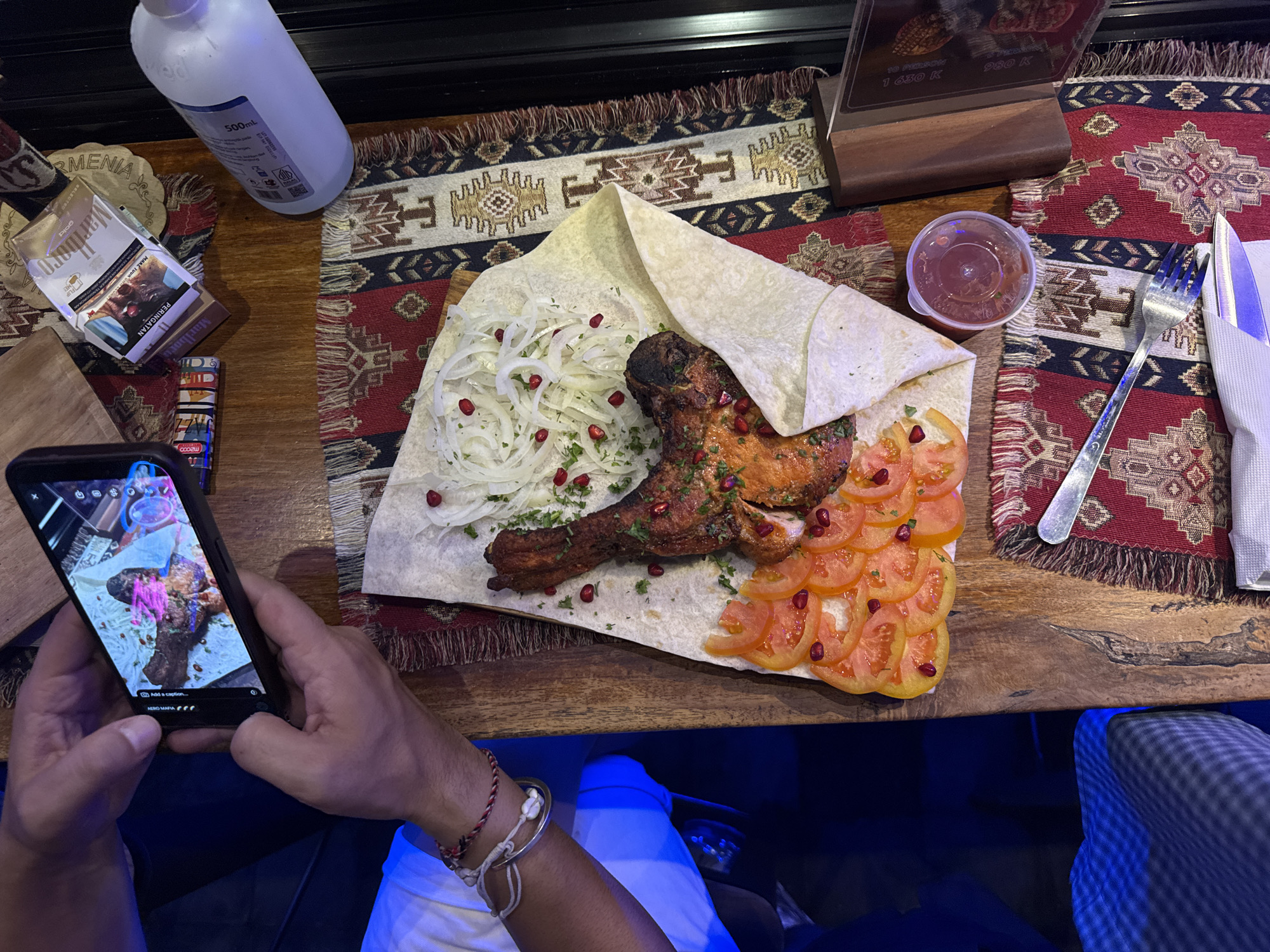The image shows an overhead view of a wooden table with a colorful rug or table runner. A plate of food is centrally located on the table. The food consists of grilled meat, sliced tomatoes, onions, herbs, and pomegranate seeds, served on a flatbread. Condiments in a clear plastic container, along with utensils and a menu stand, are also on the table. A person is holding a smartphone, taking a photo of the food. Other items include a bottle, a pack of cigarettes, and a lighter. The person's wrist is visible, adorned with bracelets. The location might be North Kuta, Indonesia, based on the information on the cigarette box visible.