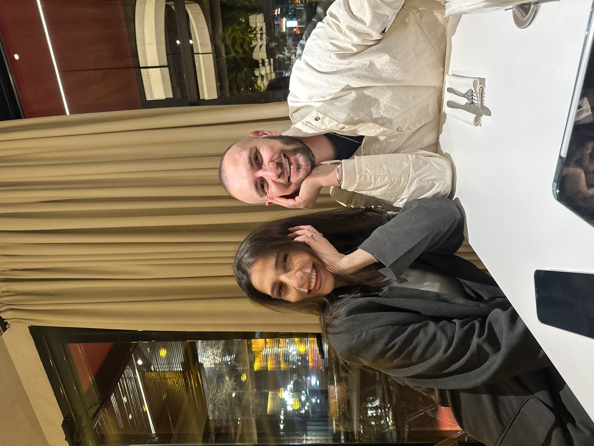 The image depicts two individuals, a female and a male, seated at a white table within an upscale restaurant or cafe in Yerevan, Armenia. The location is consistent with a modern commercial area such as Northern Avenue or near Republic Square, suggested by reflections through the large windows of palm trees and a contemporary building with curved horizontal bands and blurred urban lights.
The female, positioned on the left, has long dark brown hair and is smiling broadly, revealing her teeth. She wears a dark gray blazer over a lighter top, accessorized with a heart-shaped pendant. Her left hand, adorned with a ring on the ring finger, supports her left cheek.
The male, on the right, has a bald head and a dark stubble beard, also smiling broadly with visible teeth. He wears an off-white or cream-colored jacket over a black collared shirt, with a gold chain visible. His right hand supports his right cheek, and a silver bracelet is visible on his left wrist.
On the white table, a smartphone with a dark screen is partially visible in the foreground. To the right, a white napkin, folded around a knife and a fork, is present, along with another folded white napkin or tissue dispenser further to the right.
The background features large glass windows with dark frames, partially obscured by light beige, floor-to-ceiling drapes on the right. Reflections in the windows indicate interior warm lighting and an urban night scene outside with blurred lights and distinct architectural elements including palm trees and a building with horizontal, curved facade details. Both individuals are looking directly at the camera.