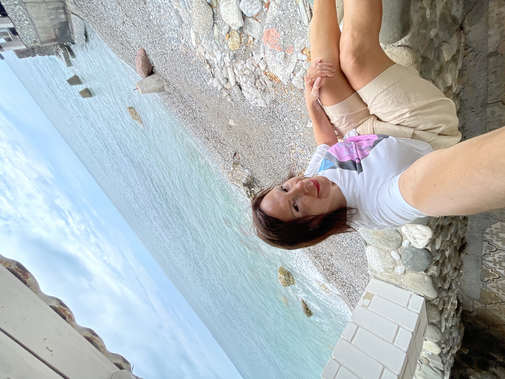 A woman with brown hair and a white t-shirt with a colorful graphic print is taking a selfie on a rocky beach. She is wearing light-colored shorts and is seated with her legs crossed. Behind her, the turquoise ocean stretches to the horizon under a cloudy sky. Concrete barriers are visible in the water and on the shore, suggesting coastal defenses or remnants of a structure. The beach is covered in small, grey pebbles and larger rocks. Part of a white, brick-like structure is in the foreground, and a textured stone wall is below it. The scene appears to be in Abaata, Abkhazia.