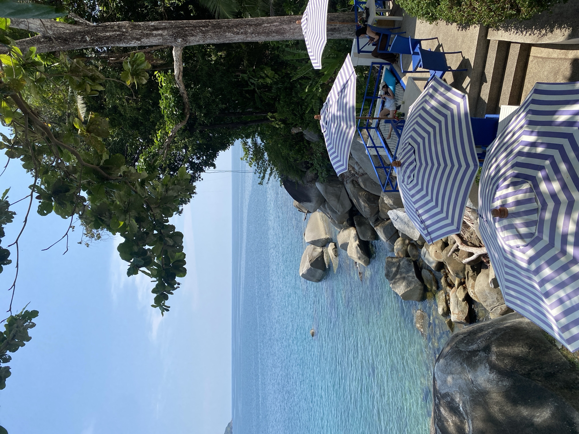 The image captures a coastal scene at Kamala, Thailand. The view is from an elevated position looking toward the sea. In the foreground, there are partially visible beach umbrellas with blue and white stripes. Below the umbrellas, rocks and pebbles form the coastline. The sea extends into the distance, with varying shades of blue. A small, light-colored object is visible in the water. On the right, there are stairs and blue chairs. On the left, tree branches and foliage are visible, contrasting with the blue sky. People are visible beneath one of the umbrellas, some seated.