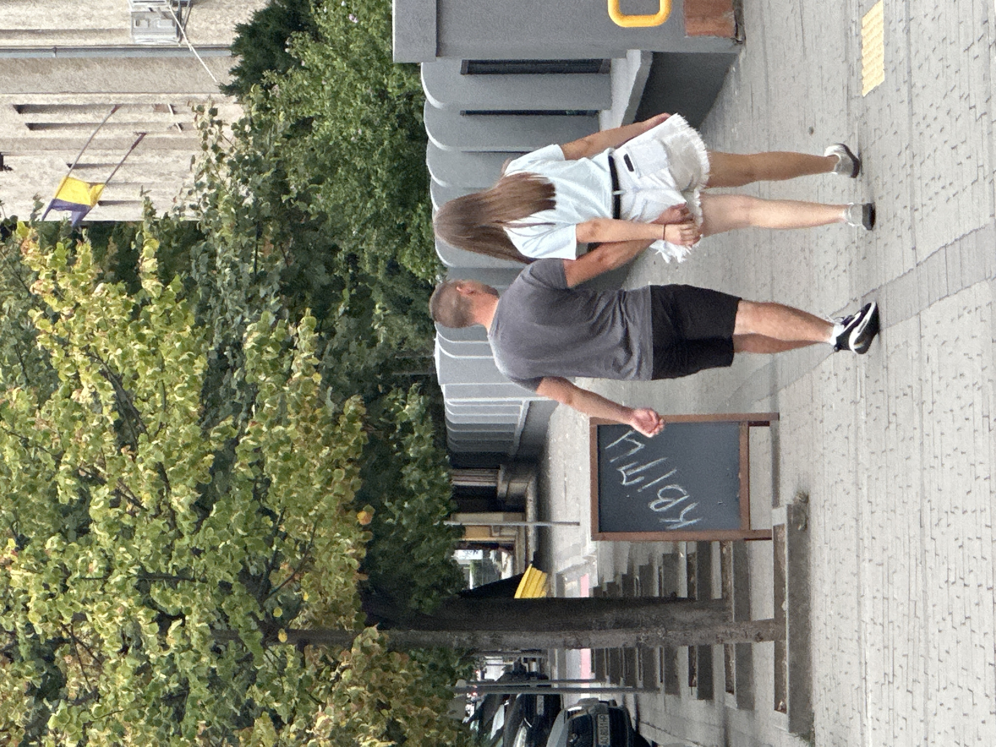 Two individuals, a male and a female, are walking hand-in-hand along a sidewalk in Uzhhorod, Ukraine. The male is wearing a grey t-shirt and black shorts. The female is wearing a light blue t-shirt and white denim shorts. They are walking away from the camera, towards the left side of the image.
To their left, a dark chalkboard sign stands on an easel, with the word "КВІТИ" (flowers in Ukrainian) written in chalk. In the background, trees with green and yellow leaves line the sidewalk, and a building with a flag flying outside is visible. Cars are parked along the street to the left of the sidewalk. The sidewalk is paved with grey interlocking bricks, and there are planters along the edge of the street. The overall environment suggests a public urban setting, possibly a street or park area. The presence of the Ukrainian flag indicates the country. The lettering on the sign, "КВІТИ", suggests a flower shop or a place selling flowers.