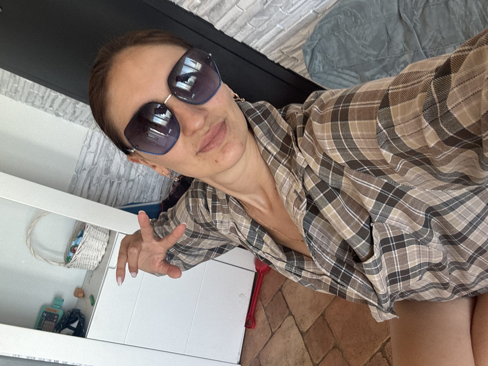 A woman with brown hair wears oversized sunglasses and a brown and white plaid shirt. She is seated indoors, with her left hand outstretched. Behind her, a white cabinet is visible with a white basket containing colorful items on a shelf. To the left of the cabinet, a red toy shovel lies on the terracotta tiled floor. The background includes a gray textured wall and a blue-green cushioned surface. The location is not specified within the image or provided in the context.