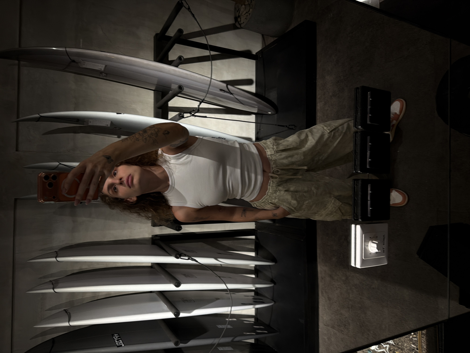 A person is taking a selfie in a store that sells surfboards. The person is wearing a white tank top, light-colored cargo pants, and orange and white sneakers. They have tattoos on their arm and hand. The store appears to be dimly lit, with surfboards displayed on racks in the background. The floor is made of concrete. In the foreground, a mirror reflects the person and the surfboards. There are also some electronic devices visible in the reflection on the floor. The location is specified as North Kuta, Indonesia.