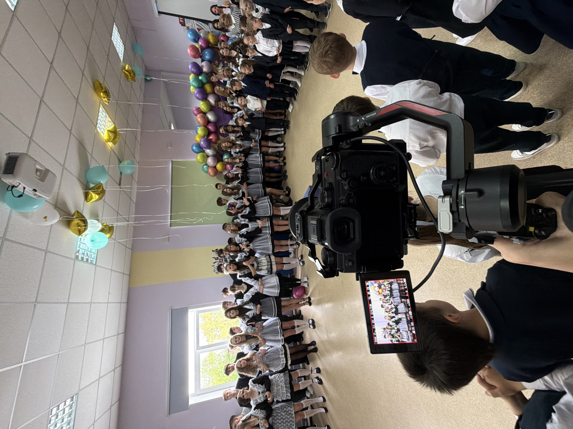 A group of schoolchildren in Krasnoyarsk, Russia, are posed for a photo or video. They are lined up against a wall, some with balloons. A person is filming them with a camera on a gimbal.