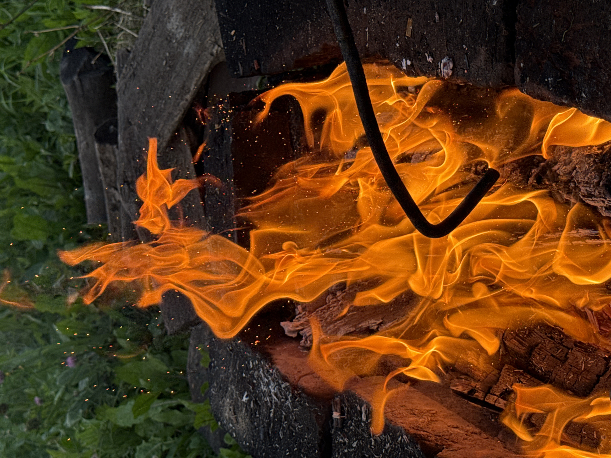 A close-up, outdoor, eye-level shot shows an active campfire with bright orange flames and glowing embers. A dark metal poker is partially visible, resting on the edge of the fire pit, suggesting recent or ongoing activity. The fire pit appears to be constructed from dark stones or bricks, with some charred wood visible within. In the background, out of focus, is lush green foliage, possibly indicating a natural setting or garden area. The lighting is dominated by the warm glow of the fire, creating a dramatic contrast with the darker surroundings and the verdant background. The image captures the raw energy and visual appeal of a roaring fire.