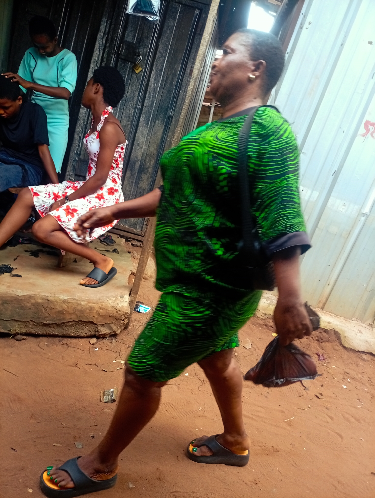 This media file captures a scene in Nkpor, Nigeria, likely outdoors on a dusty, reddish-brown ground. The primary subjects are three women.
In the foreground, a woman with dark skin walks away from the camera, her body angled to the right. She wears a vibrant green dress with a black, wavy pattern. A black strap from a bag is visible across her shoulder. She is holding a dark-colored bundle in her right hand. Her feet are adorned with dark sandals and bright green nail polish.
Behind her, another woman sits on a low concrete step. She wears a white dress with a red floral pattern. Her hair is pulled back. She looks towards the woman in the green dress.
Further behind, a third woman, also with dark skin, stands and appears to be tending to the hair of someone seated to her left (out of frame). This standing woman wears a light teal, short-sleeved dress.
The background features a textured, dark metallic surface, possibly a corrugated metal wall or door, and a lighter, ribbed metal surface. The overall lighting suggests it is daytime, and the ground texture implies a warm climate with dry conditions. No visible text is present in the image. The expressions of the women are difficult to discern clearly due to the image quality and angles, but the woman in the green dress seems to be mid-stride with her mouth open, perhaps speaking.