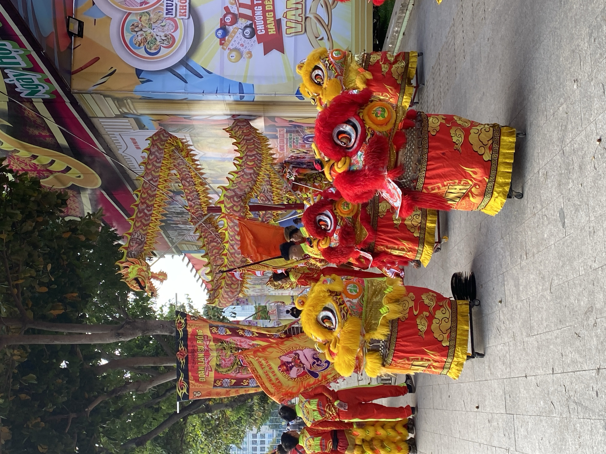 In Nha Trang Ward, Vietnam, an outdoor street scene unfolds, featuring a vibrant procession. At the forefront, several individuals are dressed in elaborate lion costumes, their heads adorned with bright red and yellow fur, large expressive eyes, and open mouths. These costumes are supported by wheels, allowing for easy movement. The lion costumes are decorated with intricate patterns of clouds and geometric designs in red and yellow, with fringed yellow skirts. Behind them, other performers wear matching red and yellow outfits, some carrying large, colorful banners with Vietnamese text. A long, segmented dragon decoration, primarily in red and yellow with green accents, is being paraded, its body arching over the procession. The backdrop consists of a large, colorful mural or banner depicting buildings, and text that appears to be related to an event or festival. Trees with green foliage line the left side of the scene, and the ground is paved with grey tiles. The atmosphere suggests a cultural celebration or performance.