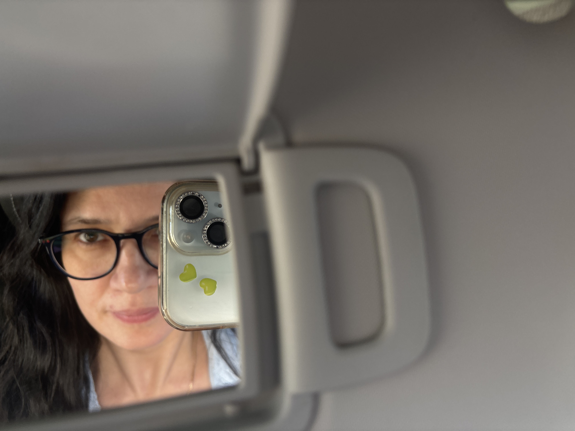 The image shows a woman with dark, wavy hair and glasses reflected in a car visor mirror. She is holding up a smartphone with a clear case adorned with rhinestones around the camera lenses and two small green heart stickers. The visor itself is gray. No location or specific details related to the city of Chaikovsky, Russia, are discernible from this image.
