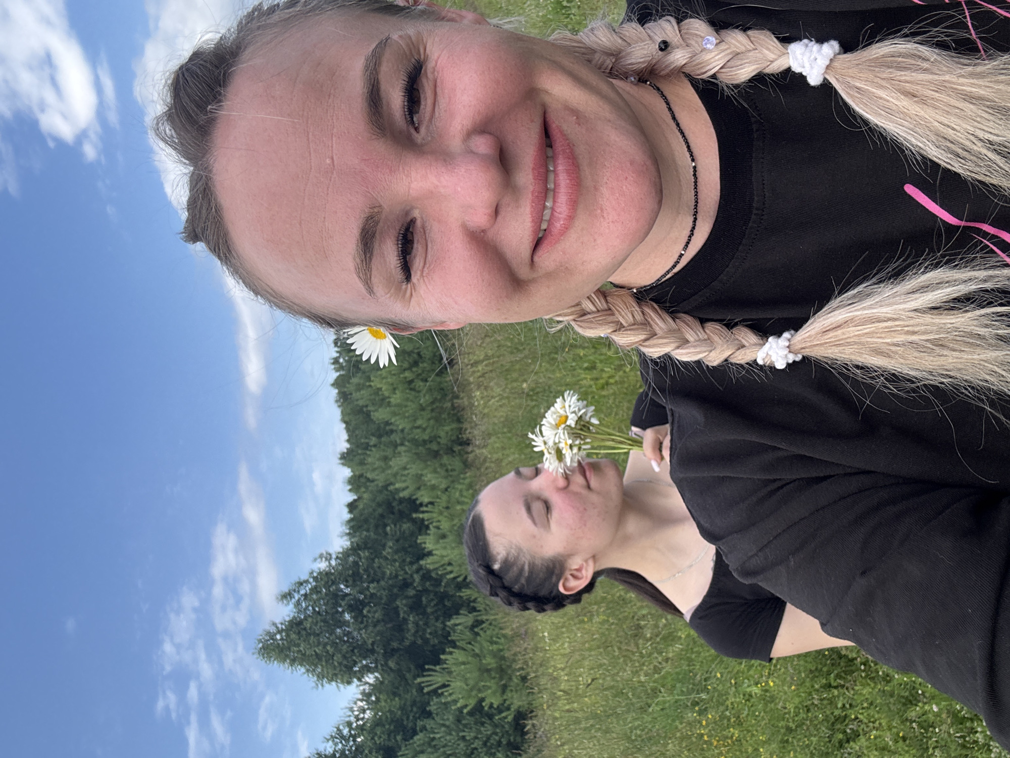 The image shows two women in a field with trees in the background and a blue sky with scattered clouds above. The woman in the foreground is taking a selfie and smiling at the camera. She has blonde hair in two braids and a daisy tucked behind her ear. The woman in the background is holding a bouquet of daisies and smelling them with her eyes closed. She has dark hair in a braid. The location appears to be a rural or natural area, possibly a meadow or field on the outskirts of Pozdyshki, Russia.
