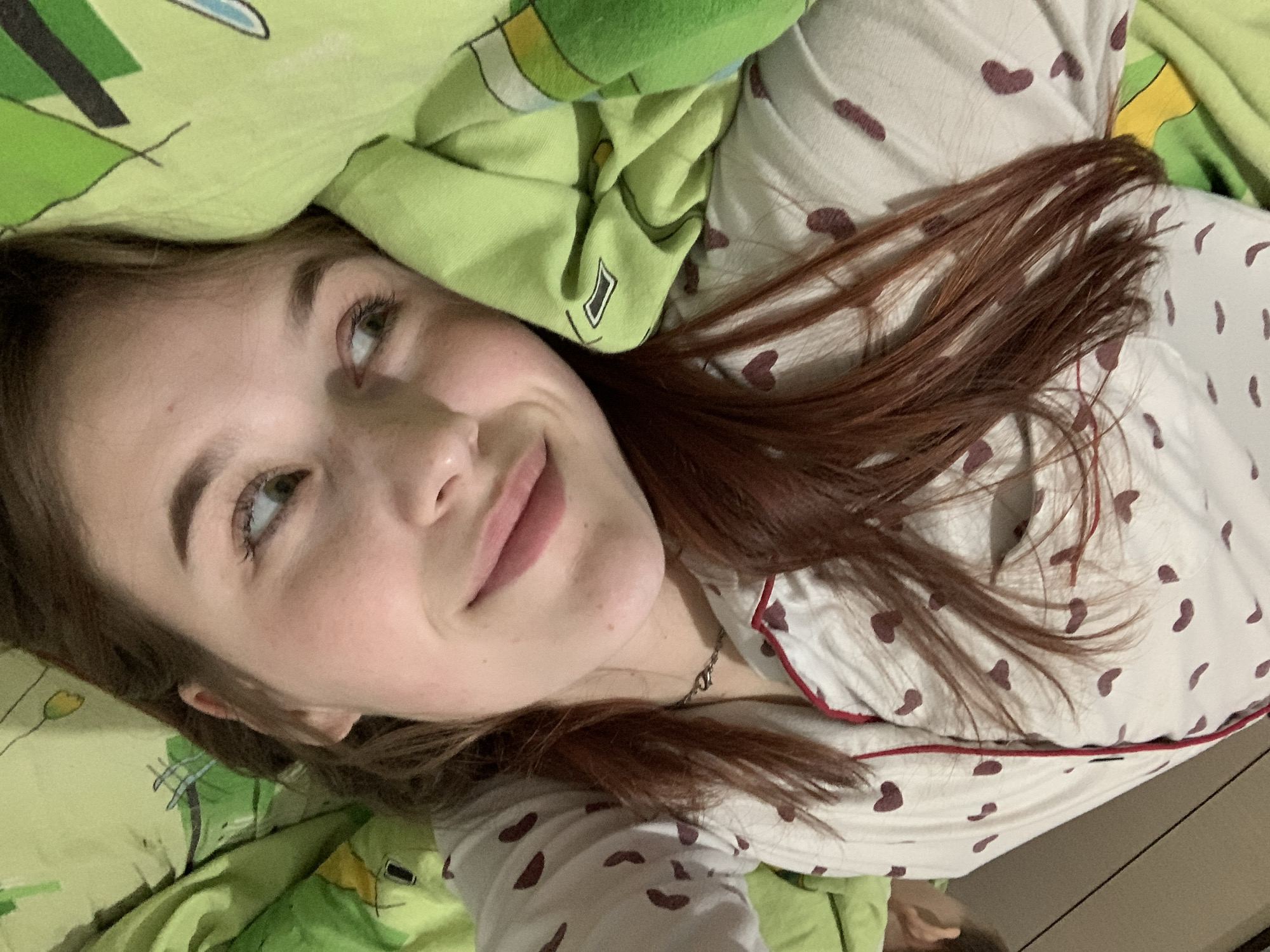 A young woman with light skin, auburn hair, and green eyes is lying down, presumably in bed, given the presence of green patterned bedding and her heart-patterned pajamas with red trim. She is looking upwards with a slight smile. A silver necklace is visible around her neck. The location in the city of Фоки, Russia is not discernible from this image.
