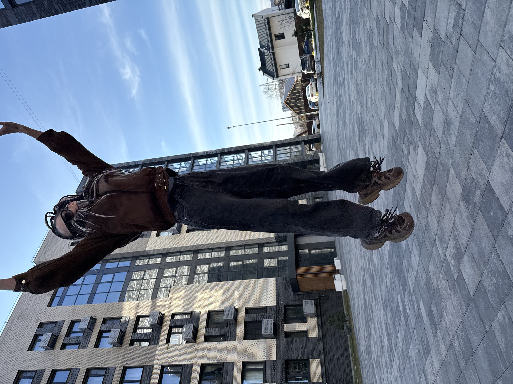 A person is posing in front of modern buildings and a brick-paved ground. The person is wearing a brown jacket, dark jeans, and sneakers. The buildings feature large windows and a mix of light and dark brick. In the background, a blue sky with light clouds is visible, along with a residential area. The photo seems to be taken on a street in Rusanovo, Russia.