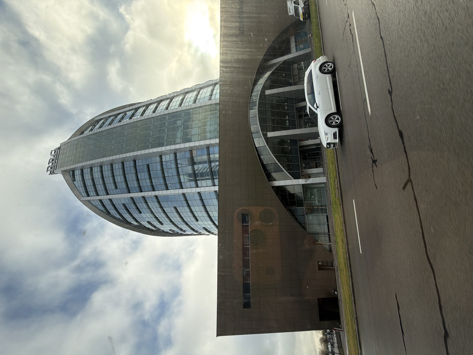 The image depicts a view in Sofia, Bulgaria, featuring the Capital Fort building complex. The distinctive rounded glass-facade tower, marked with "BCC", stands prominently against a sky with clouds. Adjacent to the tower is a modern building with a curved glass facade. A white car is parked on a roadway alongside the building. The street is paved and has painted white lane markers. The scene conveys a contemporary urban environment.