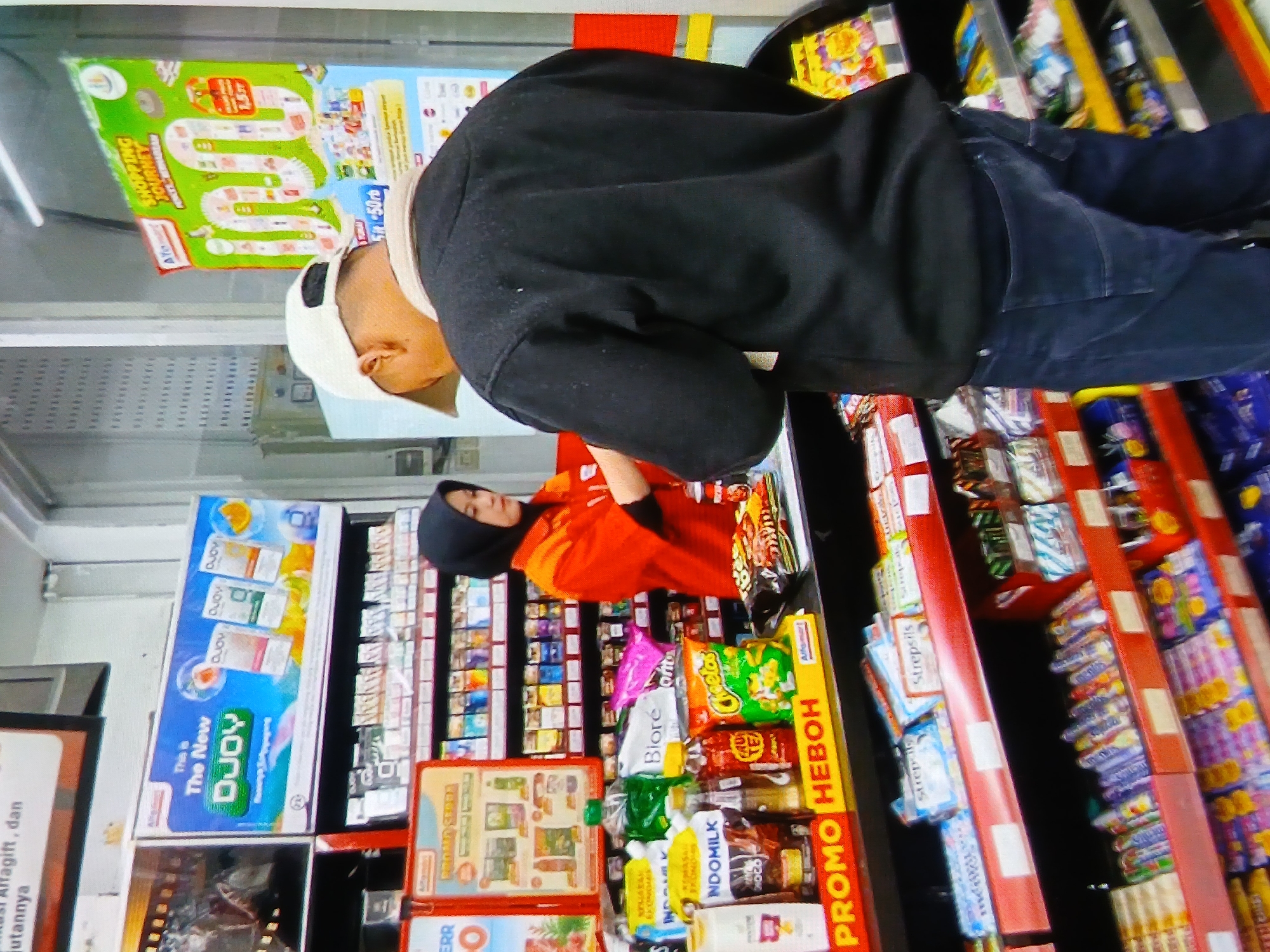The image depicts the interior of an Alfamart convenience store, likely located in Setia Bakti, Indonesia, given the contextual clues of store branding and product packaging in Indonesian. In the foreground, a customer wearing a black sweatshirt and a white baseball cap is standing at the counter, facing away from the camera. A female cashier, wearing an orange uniform and a black hijab, stands behind the counter, interacting with the customer.
The counter is laden with various convenience store items, including bagged snacks like Cheetos, bottled drinks, and boxes of Indomilk. Prominent signage indicates "PROMO HEBOH" (Exciting Promo) and "PROMO SERU" (Fun Promo). Behind the counter, shelves are stocked with a variety of packaged goods, including health and beauty products and snacks.
In the background, an advertisement for "The New DJOY" is displayed, featuring product packaging. Another large green promotional poster titled "SHOPPING JOURNEY" is also visible, illustrating a path with various store items. The lighting suggests an indoor environment with fluorescent overhead lights. The overall scene captures a typical transaction at an Indonesian convenience store.