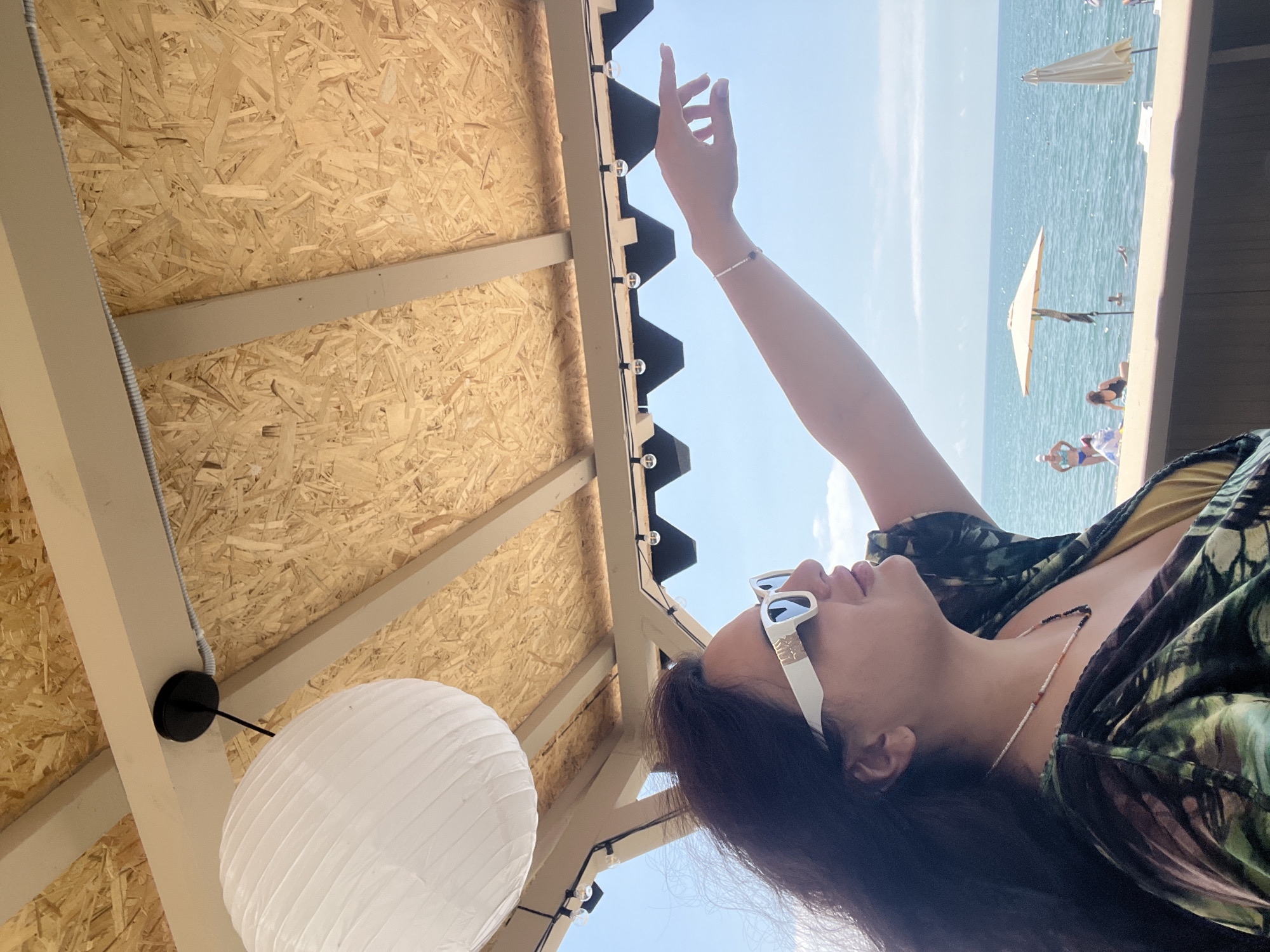 The image depicts a woman with dark hair wearing sunglasses and a colorful patterned top, seated outdoors. She is looking to her right and pointing with her right arm, which is adorned with a bracelet. Behind her, the sea is visible with several people in the water and on the beach, along with beach umbrellas. The foreground features a wooden structure with a slatted ceiling and a hanging lantern. The setting appears to be a beachside establishment in Sokhumi, Abkhazia, suggesting a relaxed, recreational atmosphere.