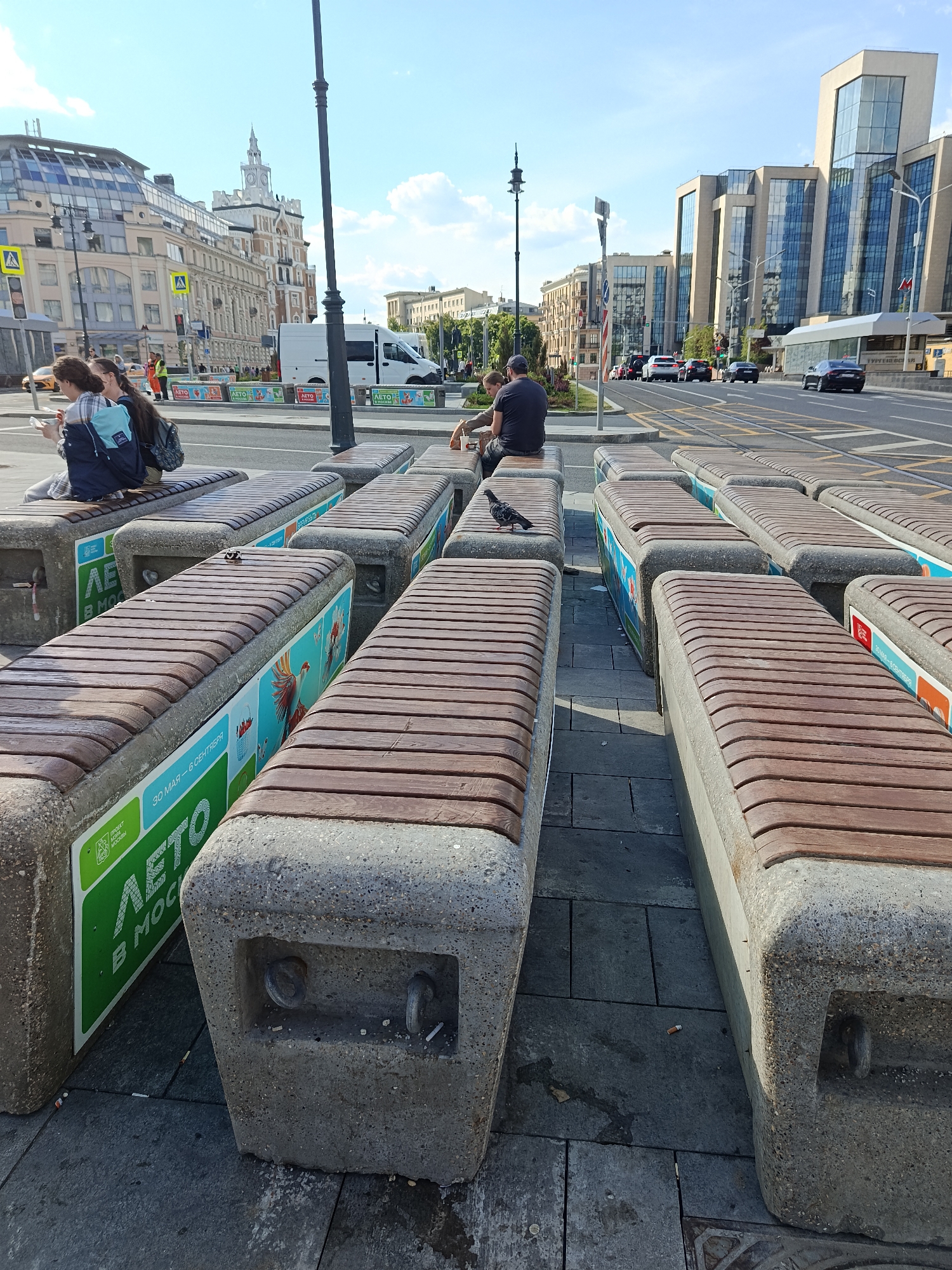 This image depicts an urban street scene on a bright, sunny day, likely in Moscow, Russia, as indicated by the Cyrillic text "ЛЕТО В МОСКВЕ" (Summer in Moscow) and "ПРАВИТЕЛЬСТВО МОСКВЫ" (Government of Moscow) prominently displayed on the benches. The dates "30 МАЯ - 6 СЕНТЯБРЯ" (May 30 - September 6) are also visible, suggesting a seasonal public initiative.
In the foreground, numerous concrete benches with wooden slatted tops are arranged in parallel rows, providing seating for pedestrians. Several people are relaxing on these benches. On the left, two individuals, likely women, are seated, one holding a phone or book, with a blue backpack resting beside her. Further along the row of benches, a man in a dark t-shirt and cap sits with another person, both facing away from the camera. A pigeon is perched on one of the unoccupied benches in the middle ground.
Beyond the seating area lies a wide, multi-lane road with cars and a white van traversing it. On either side of the street, a blend of architectural styles is apparent; to the left, an older building with a distinctive clock tower stands alongside other structures, while to the right, modern buildings with extensive glass facades rise against the sky. The sky is clear blue with scattered white clouds, suggesting pleasant weather.
Other visible details include streetlights, traffic signals, and street markings on the asphalt. The concrete bases of some benches feature recessed areas containing cigarette butts, indicating their use as impromptu ashtrays. Some benches also carry colorful advertisements with artistic illustrations, such as a large bird or berries, reinforcing the "Summer in Moscow" theme. The scene portrays a typical bustling city environment where people pause to rest and observe the urban activity.